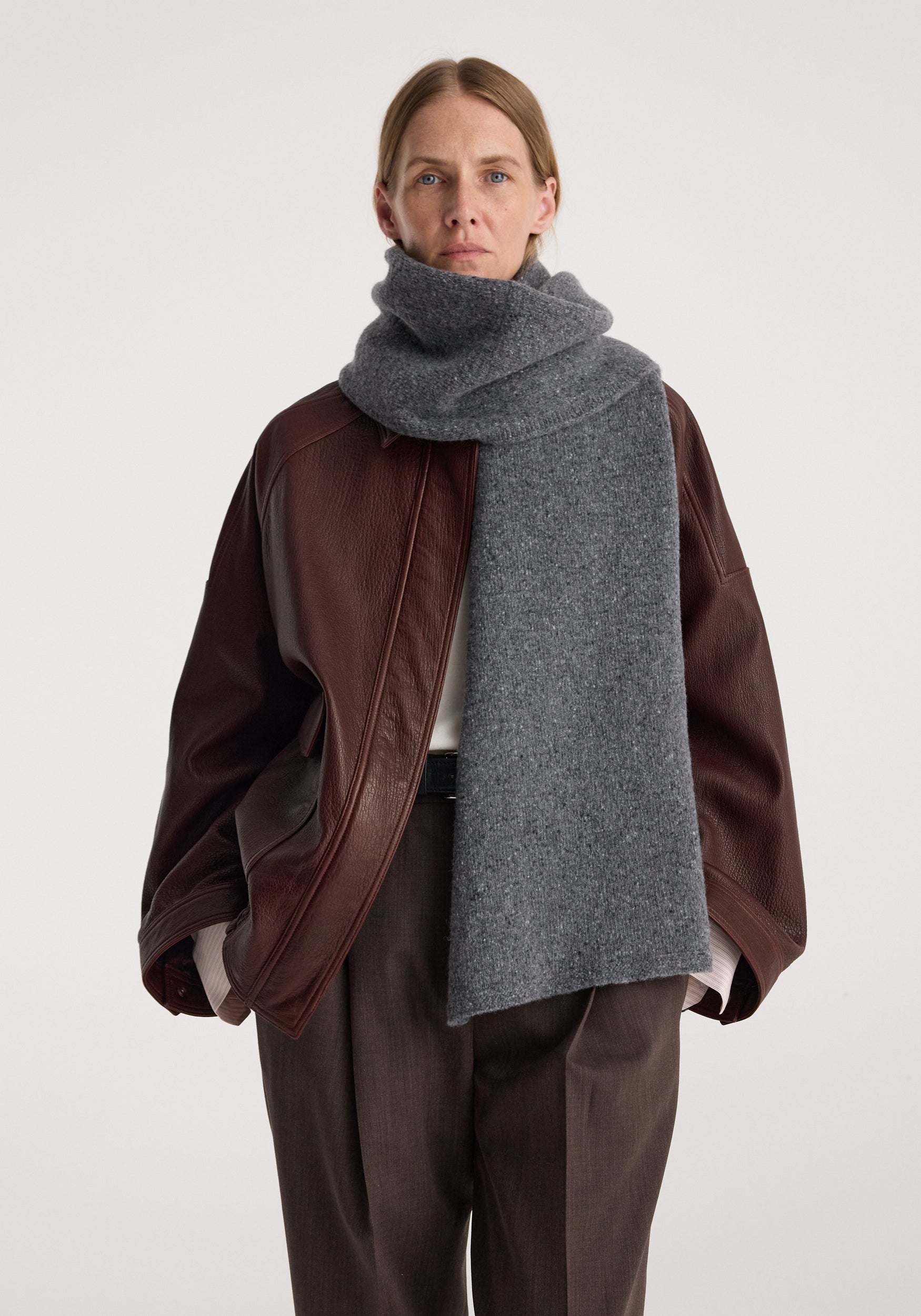 Woman wearing a thick dark gray scarf wrapped around her neck and draped over a brown leather jacket and loose brown trousers, showcasing the Knitted mouliné scarf | anthracite melange.