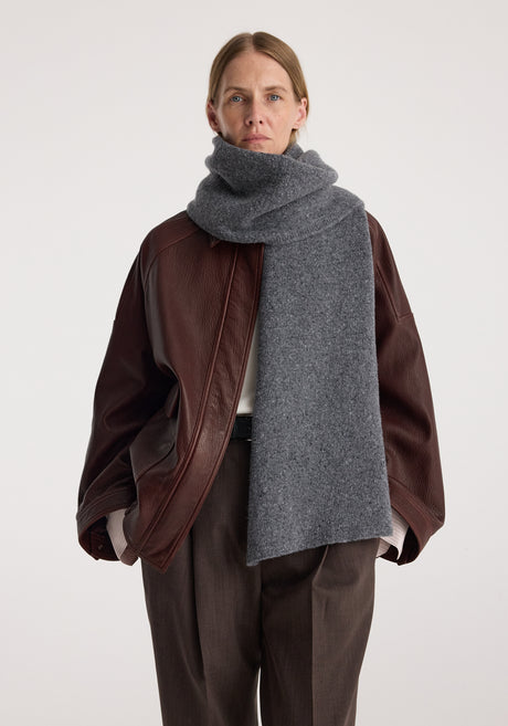 Woman wearing a Knitted mouliné scarf | anthracite melange wrapped around her neck paired with a brown leather jacket, dark brown trousers, and black heeled sandals.