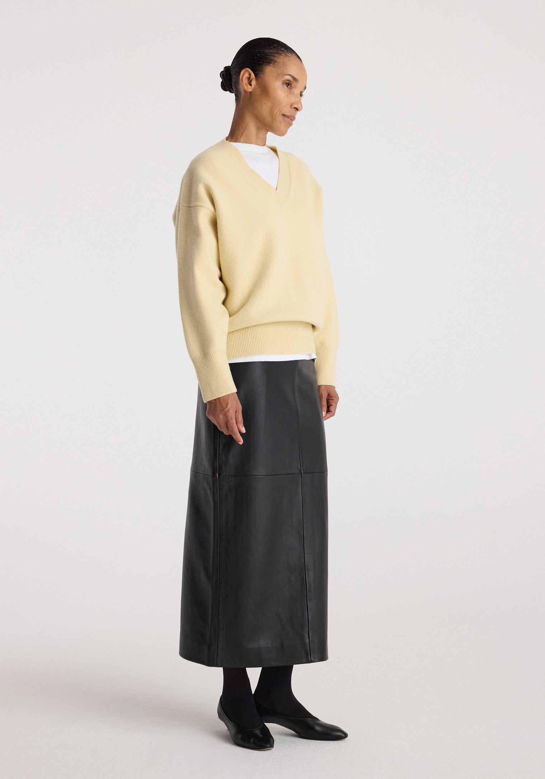 Woman wearing a Classic v-neck jumper | butter in a soft yellow shade paired with a white undershirt, black long skirt, black tights, and black flats against a plain background.