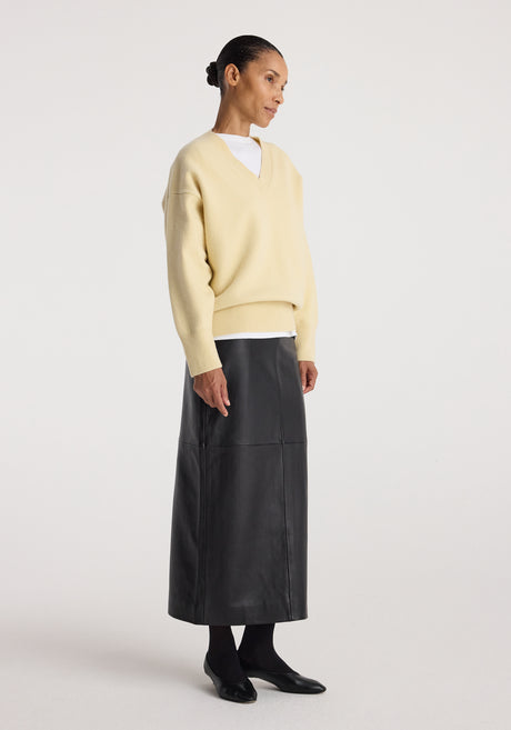 Woman wearing a Classic v-neck jumper | butter in pale yellow, paired with a long black leather skirt, black tights, and black flat shoes, standing against a plain background.