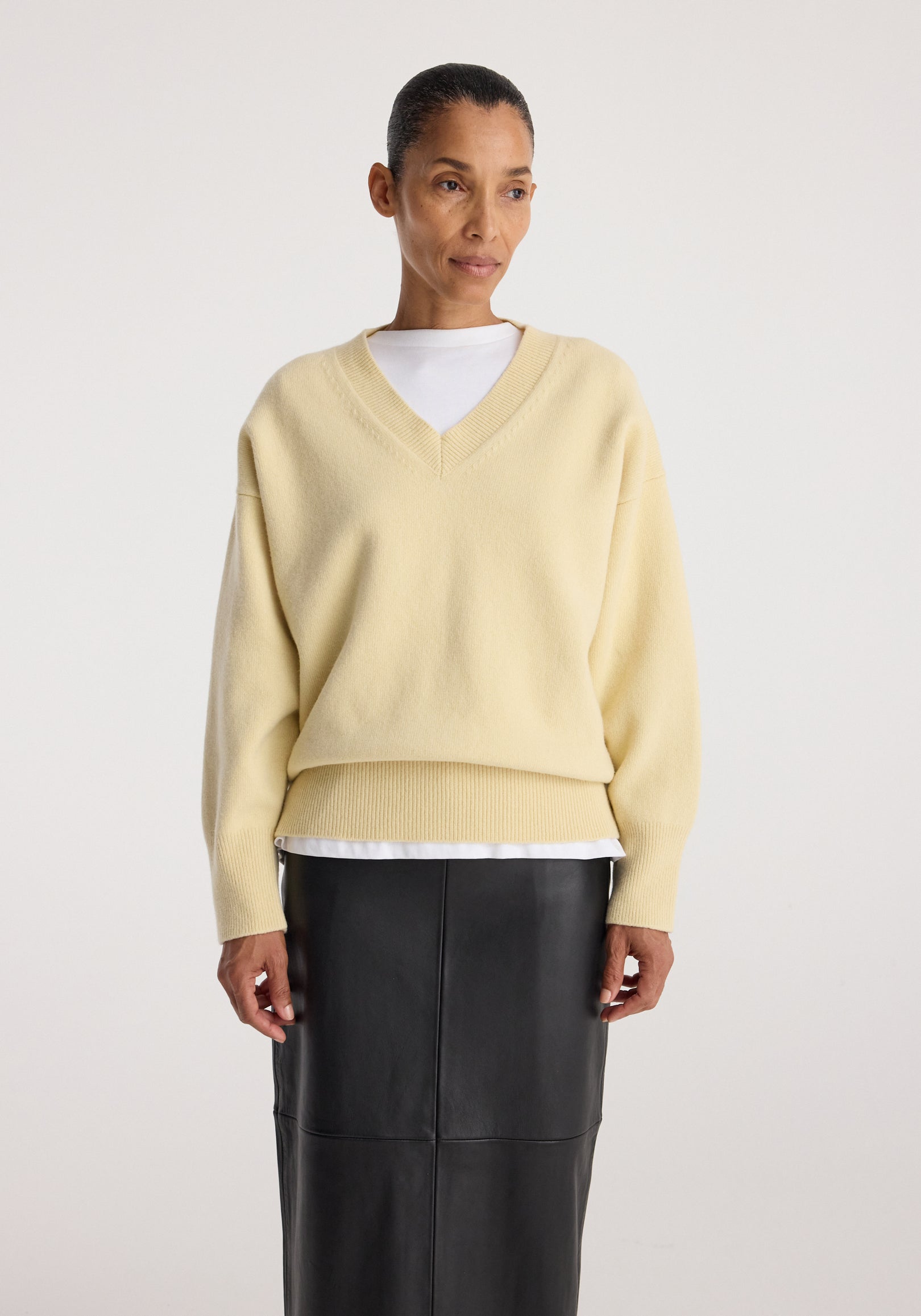 Woman wearing a Classic v-neck jumper | butter in a soft yellow shade layered over a white shirt paired with a black leather skirt against a plain background.