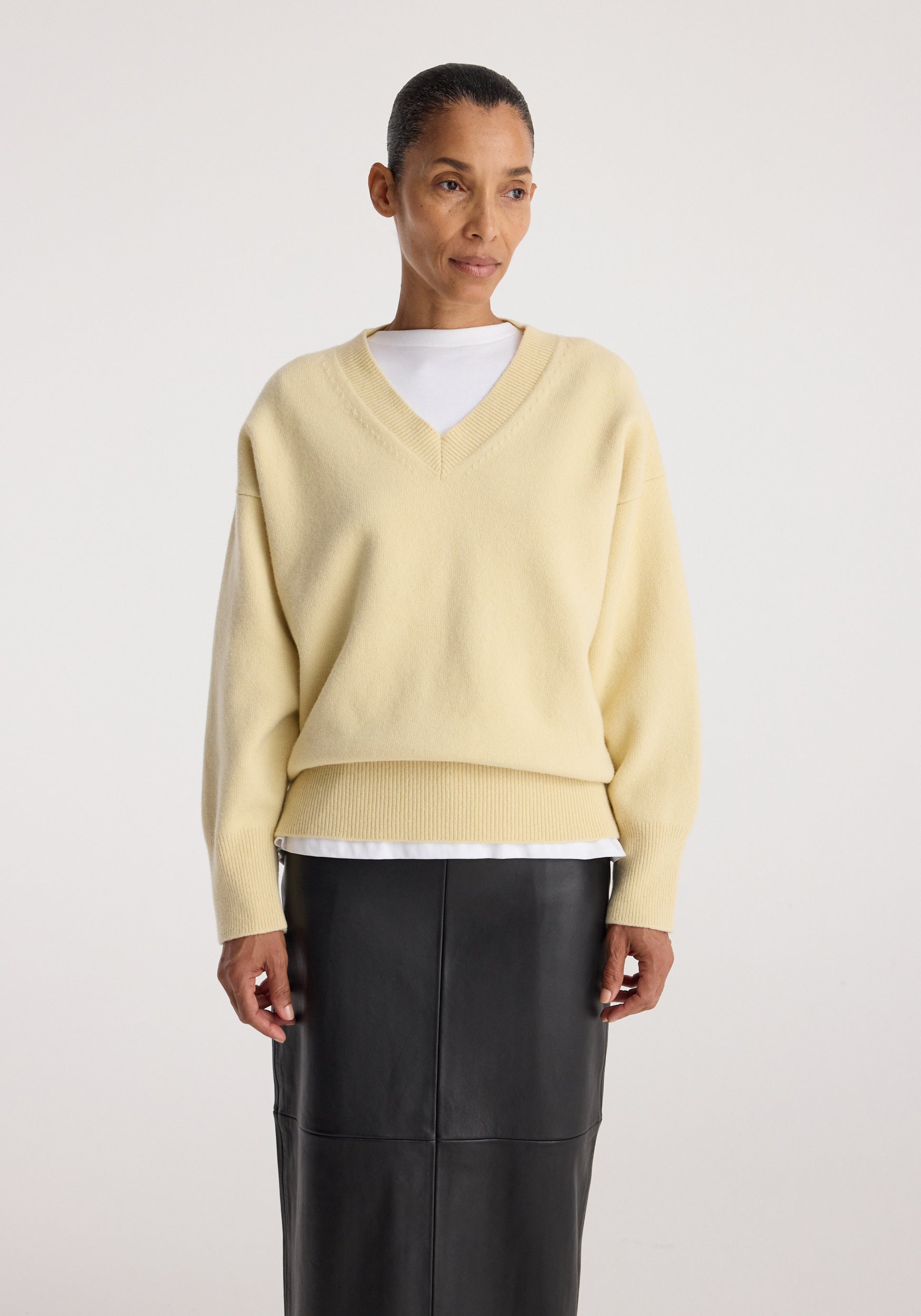 Woman wearing a Classic v-neck jumper | butter in a soft yellow shade layered over a white shirt paired with a black leather skirt against a plain background.