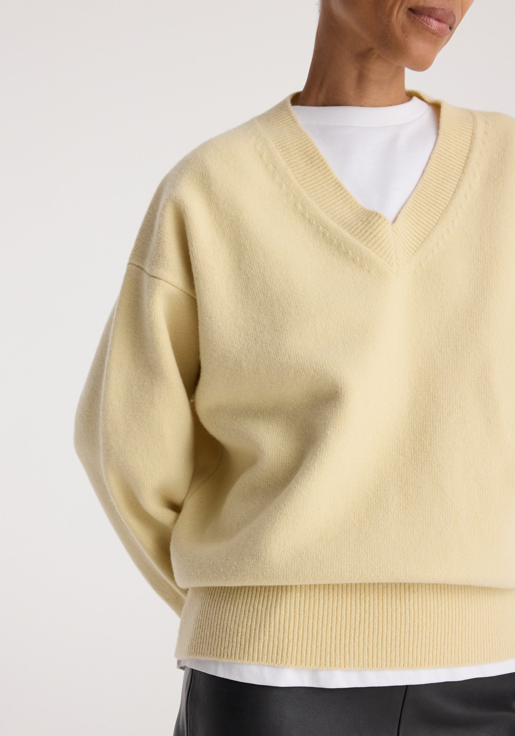Close-up of a person wearing a soft butter-colored Classic v-neck jumper | butter layered over a white shirt with black pants against a light neutral background.