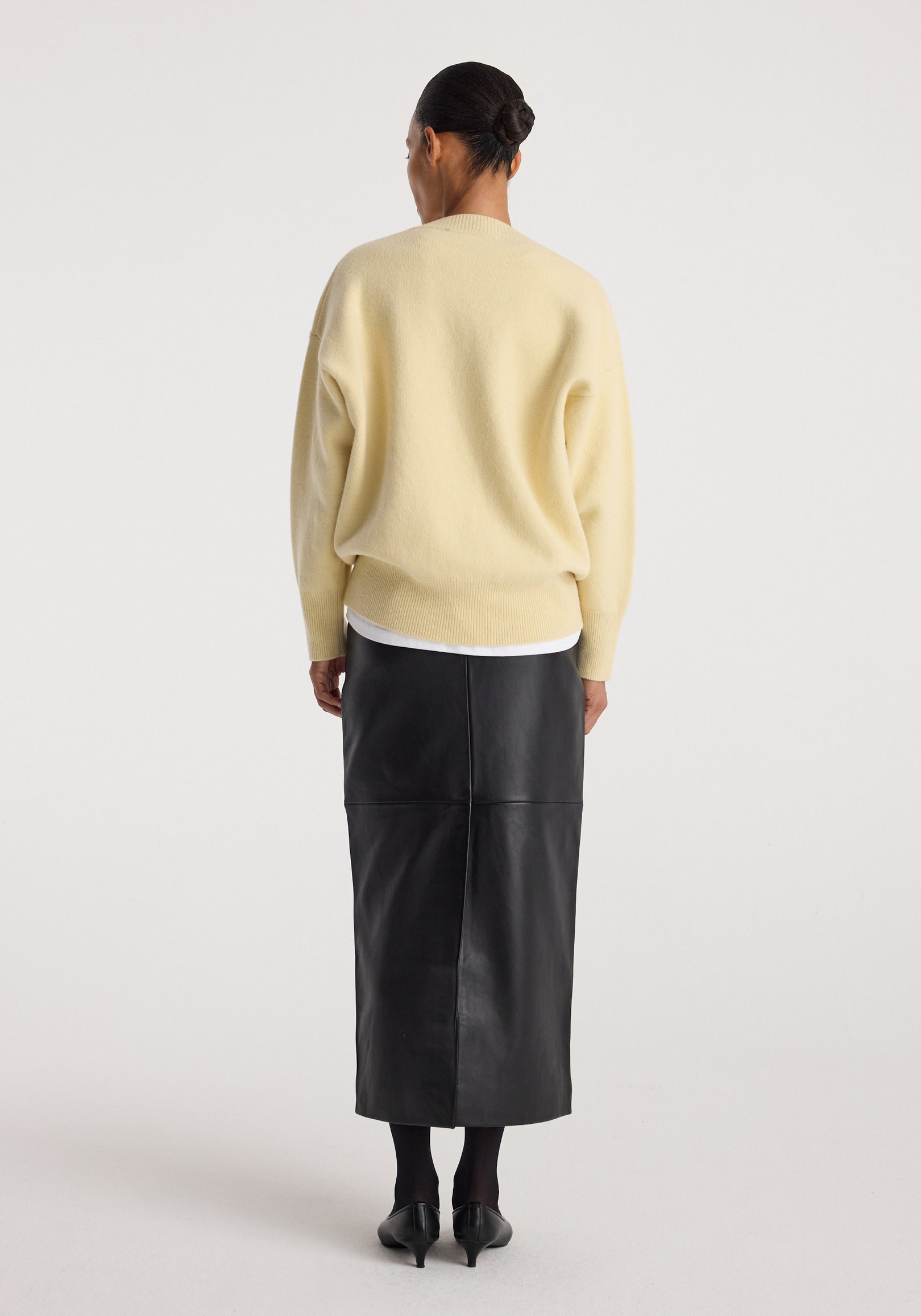 Rear view of a woman wearing a Classic v-neck jumper | butter in soft yellow, paired with a black leather midi skirt and black heels against a plain white background.