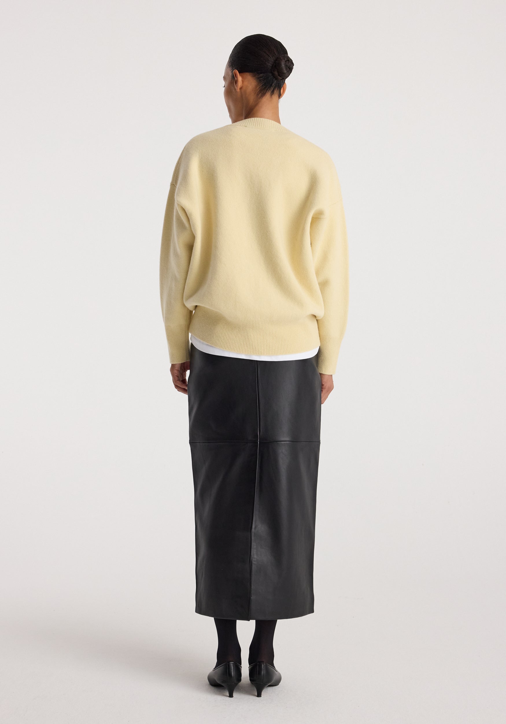 Rear view of a woman wearing a Classic v-neck jumper | butter in soft yellow, paired with a black leather midi skirt and black heels against a plain white background.