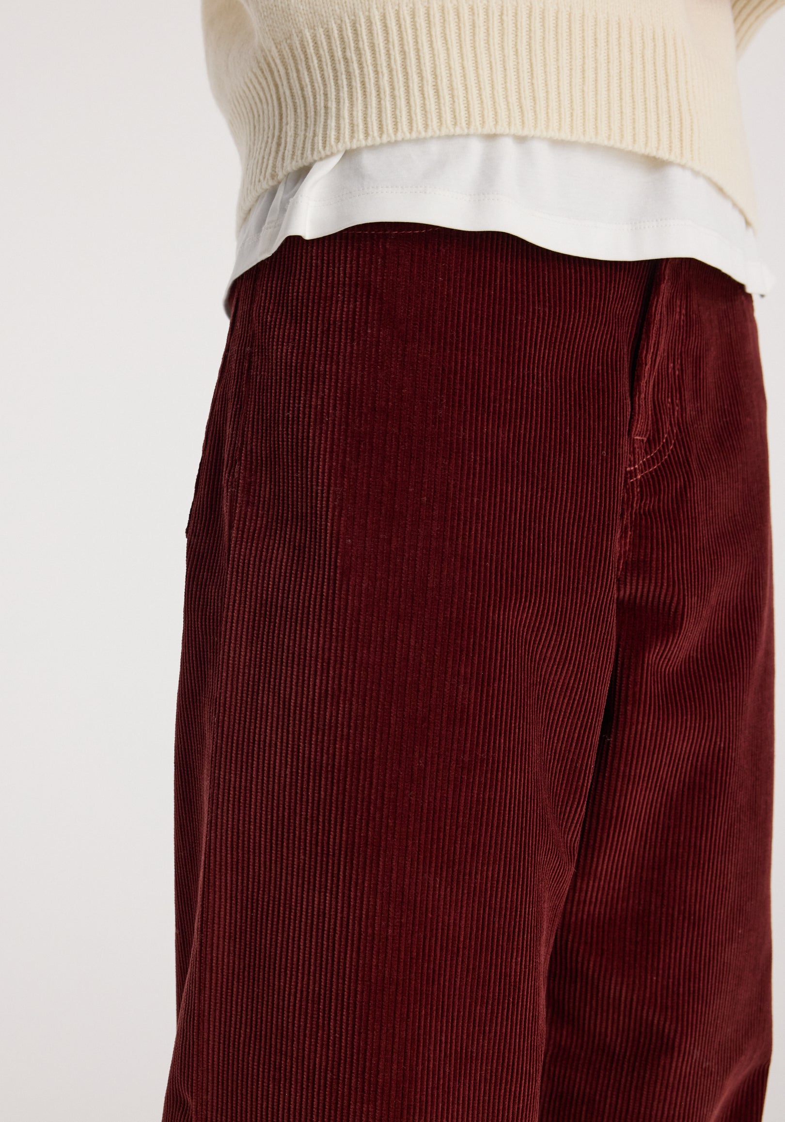 Close-up of a person wearing Bruun - Signature baggy corduroy trousers | wine in a deep red color paired with a cream knit sweater and white undershirt.