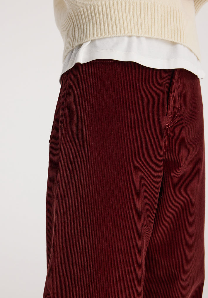 Close-up of a person wearing Bruun - Signature baggy corduroy trousers | wine in a deep red color paired with a cream knit sweater and white undershirt.