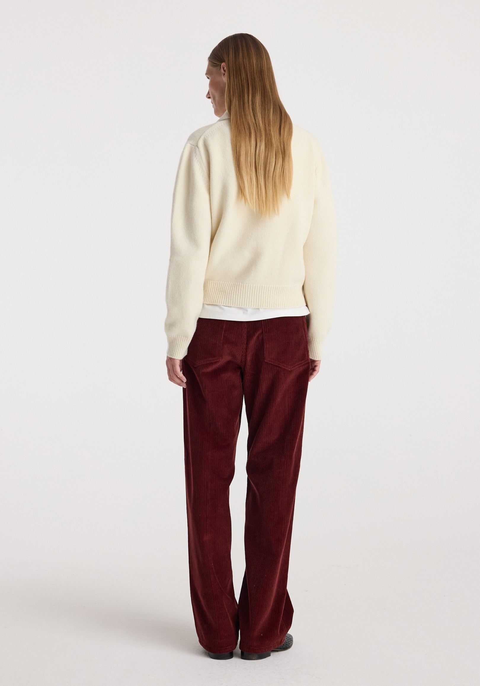 A person wearing Bruun - Signature baggy corduroy trousers | wine, styled with a cream knit sweater, standing facing away on a plain white background.