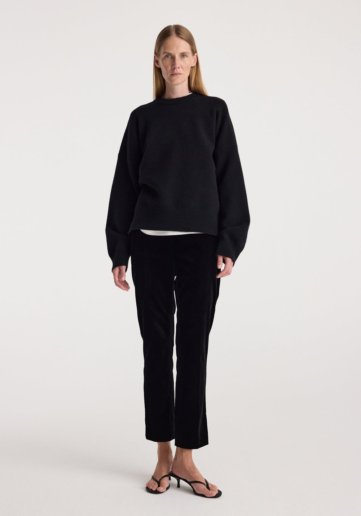 Woman wearing black Cigarette leg velvet tailored trousers | black paired with a loose black sweater and black open-toe heels, standing against a plain white background.