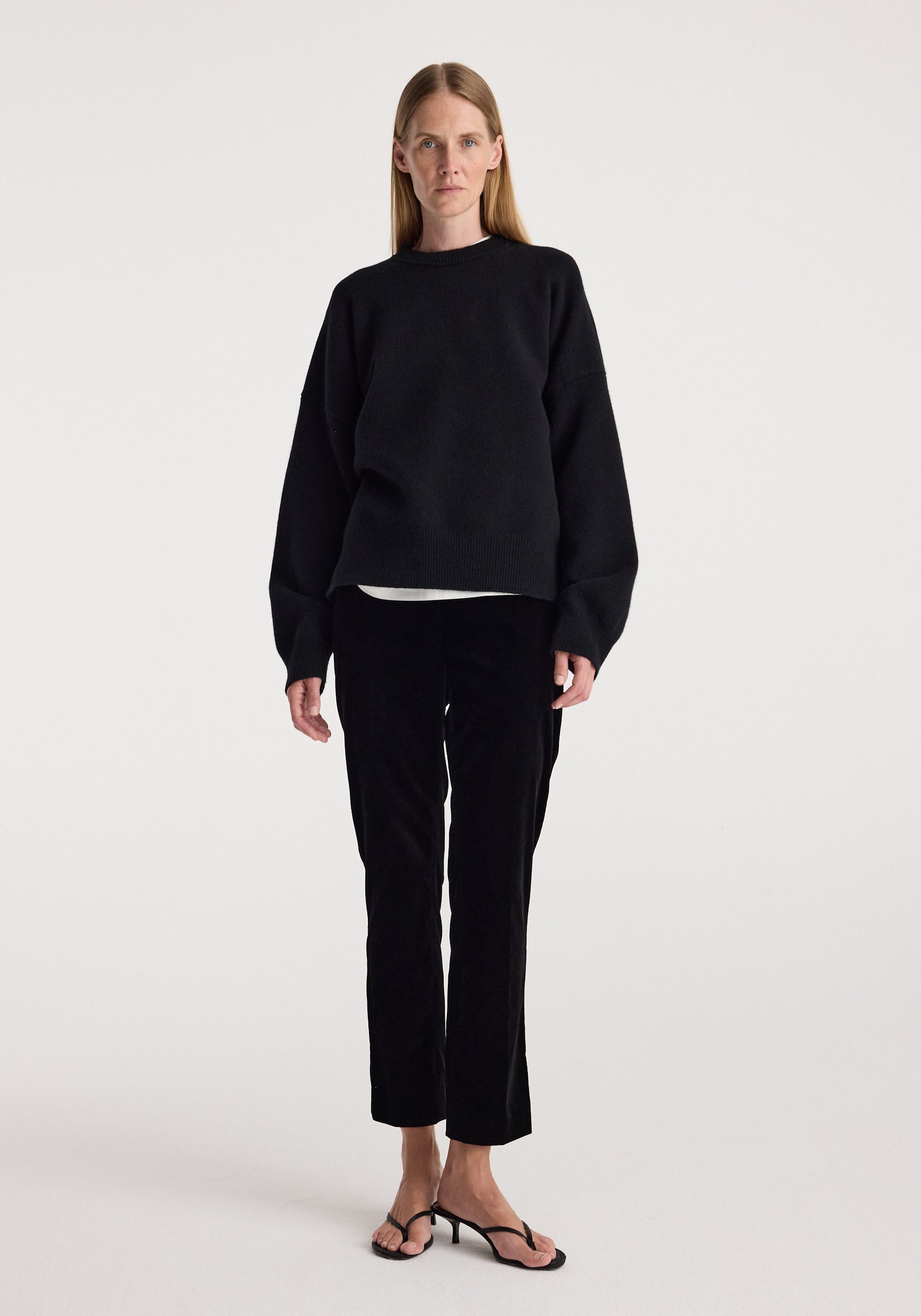 Woman wearing black Cigarette leg velvet tailored trousers | black paired with a loose black sweater and black open-toe heels, standing against a plain white background.