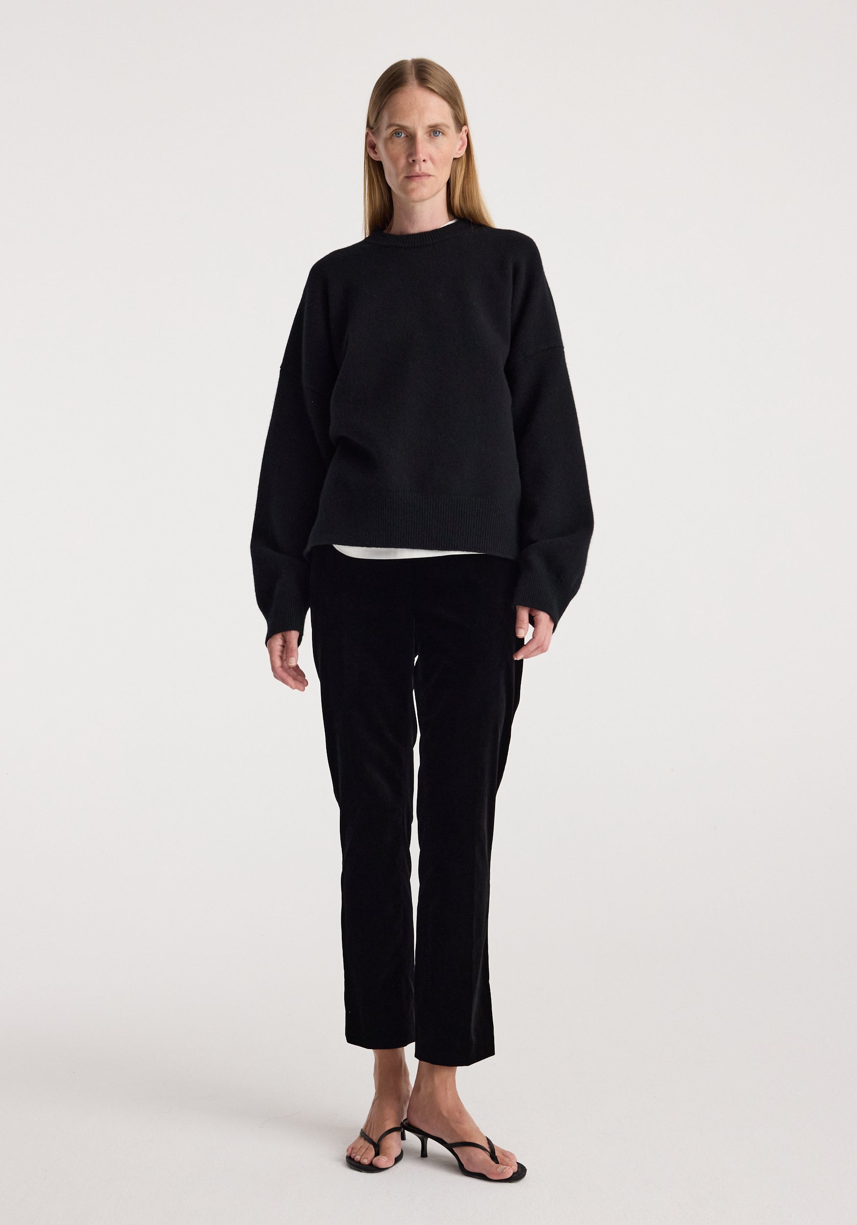 Woman wearing black Cigarette leg velvet tailored trousers | black paired with a loose black sweater and black open-toe heels, standing against a plain white background.