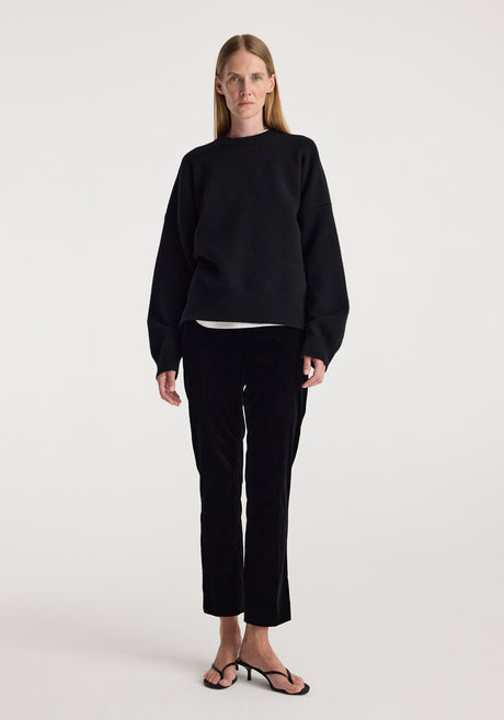 Woman wearing black Cigarette leg velvet tailored trousers | black paired with a loose black sweater and black open-toe heels, standing against a plain white background.