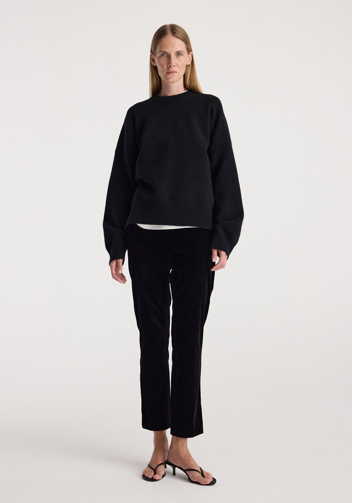 Woman wearing black Cigarette leg velvet tailored trousers | black paired with a loose black sweater and black open-toe heels, standing against a plain white background.