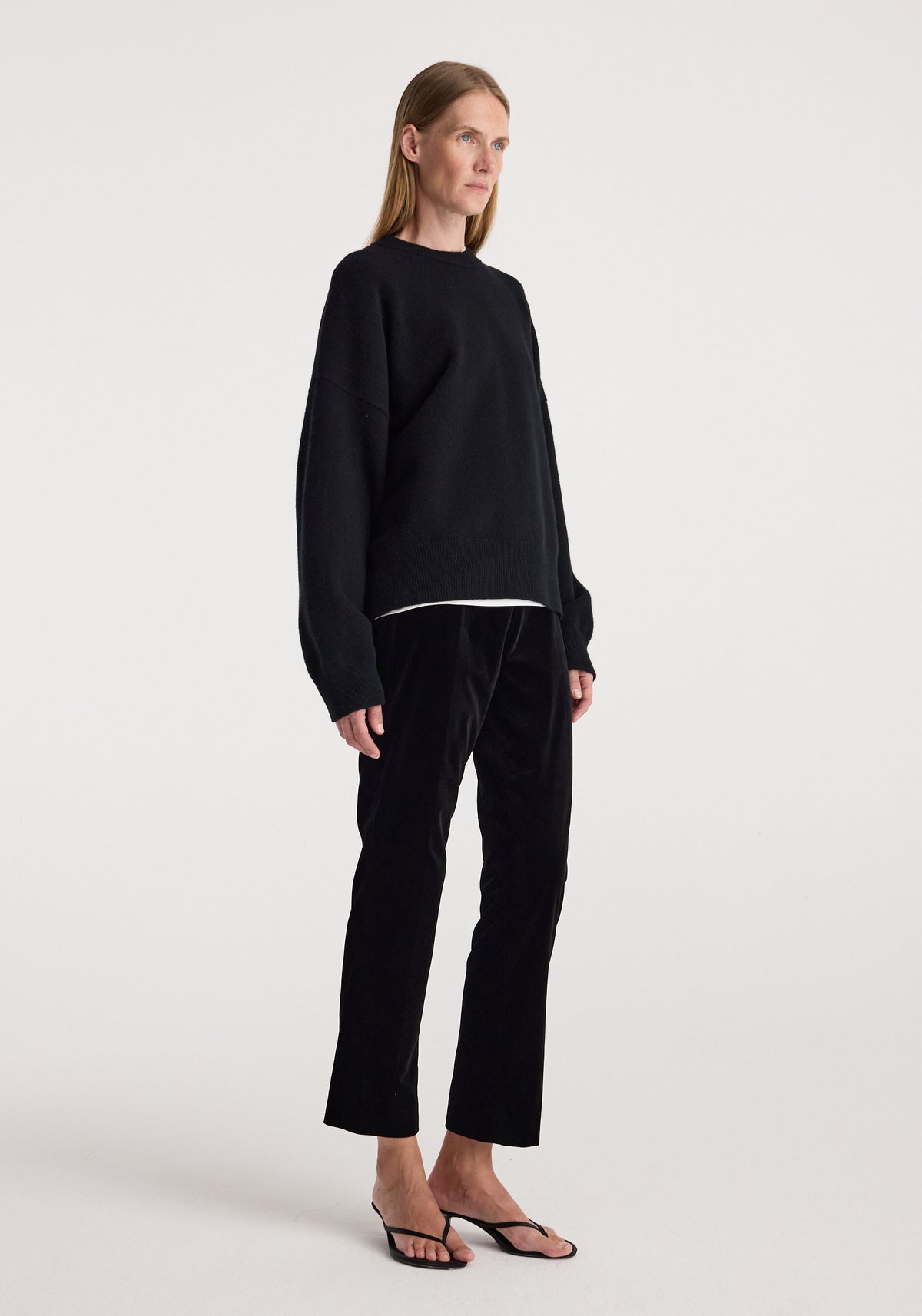 Woman standing wearing cigarette leg velvet tailored trousers | black paired with a loose black sweater and black open-toe heels against a plain background.
