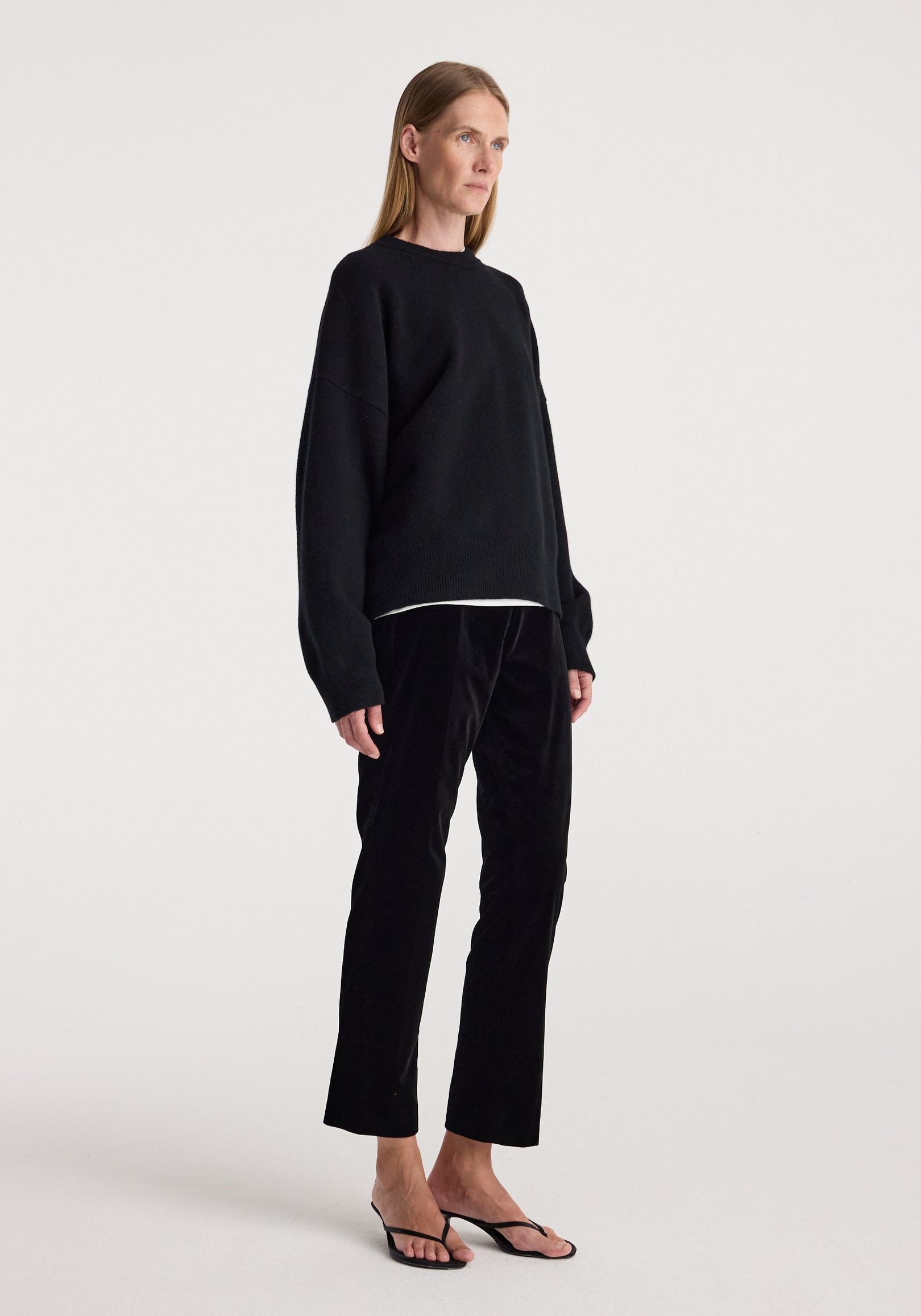 Woman standing wearing cigarette leg velvet tailored trousers | black paired with a loose black sweater and black open-toe heels against a plain background.