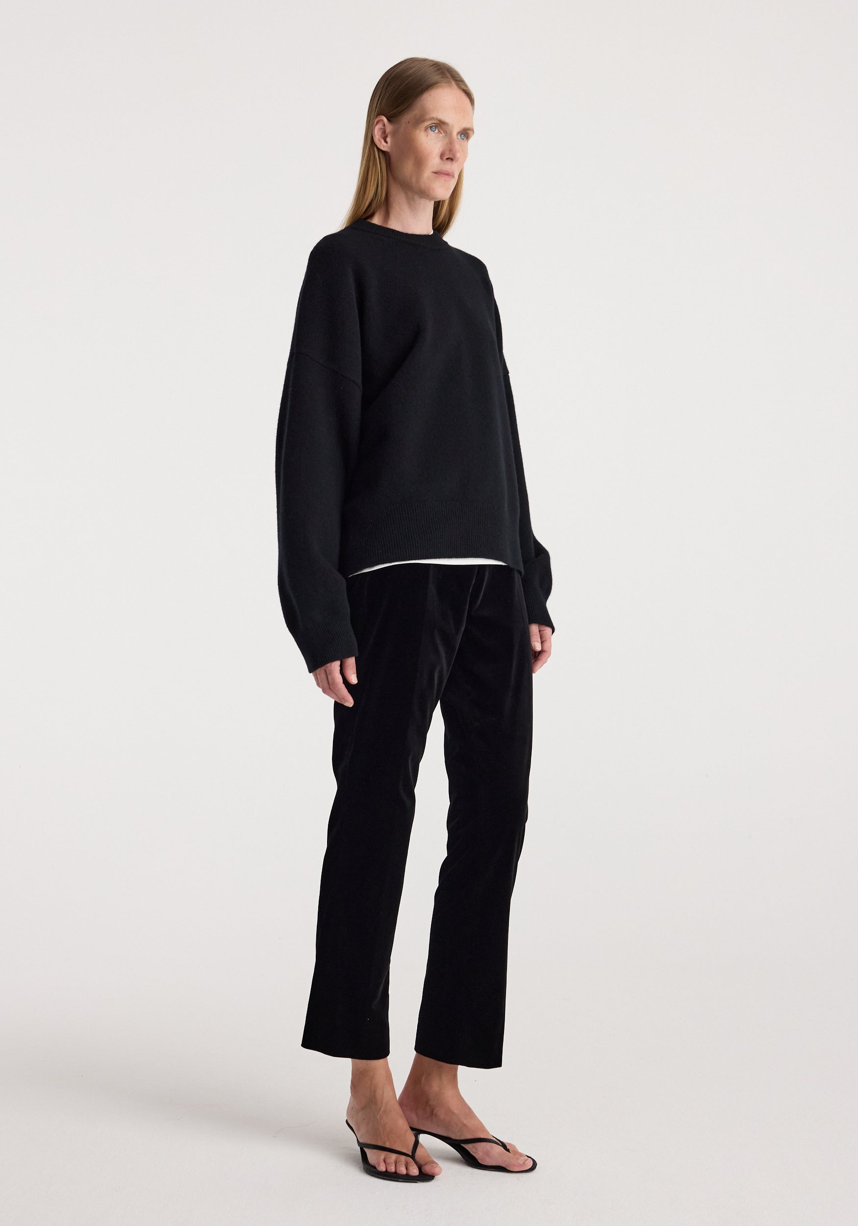 Woman standing wearing cigarette leg velvet tailored trousers | black paired with a loose black sweater and black open-toe heels against a plain background.