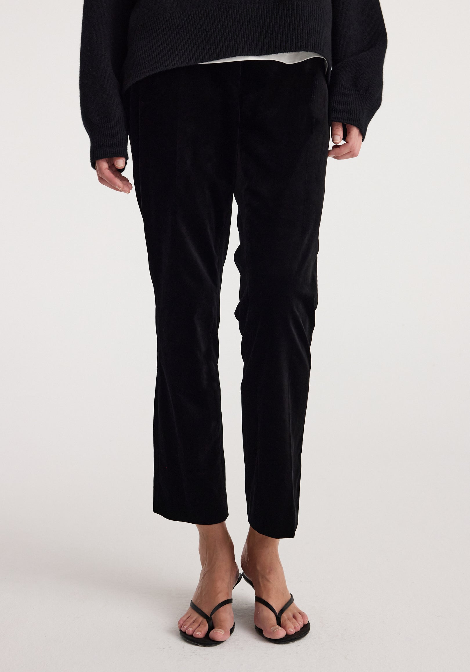 Front view of a person wearing cigarette leg velvet tailored trousers | black, styled with a black top and black open-toe sandals.