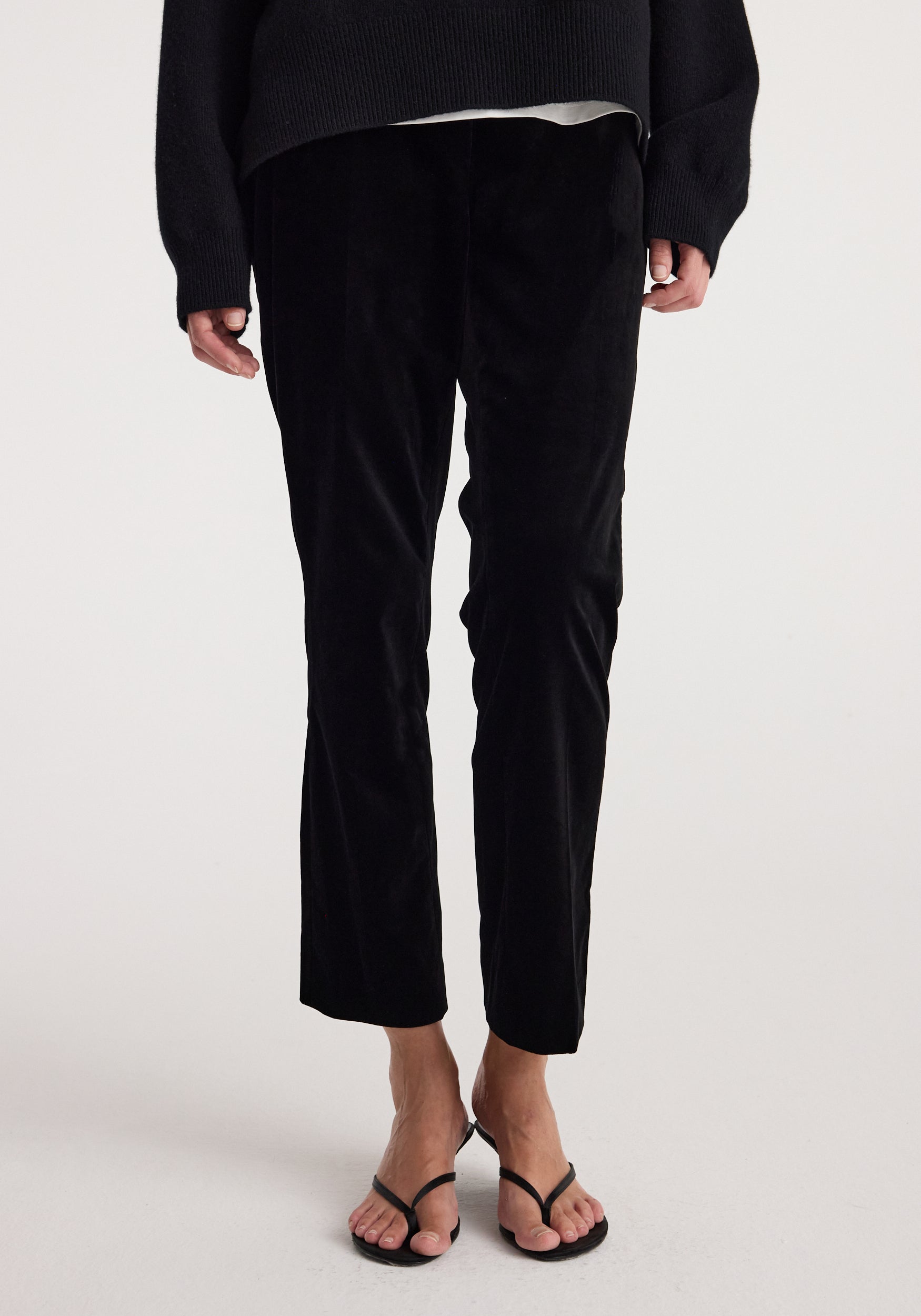 Front view of a person wearing cigarette leg velvet tailored trousers | black, styled with a black top and black open-toe sandals.