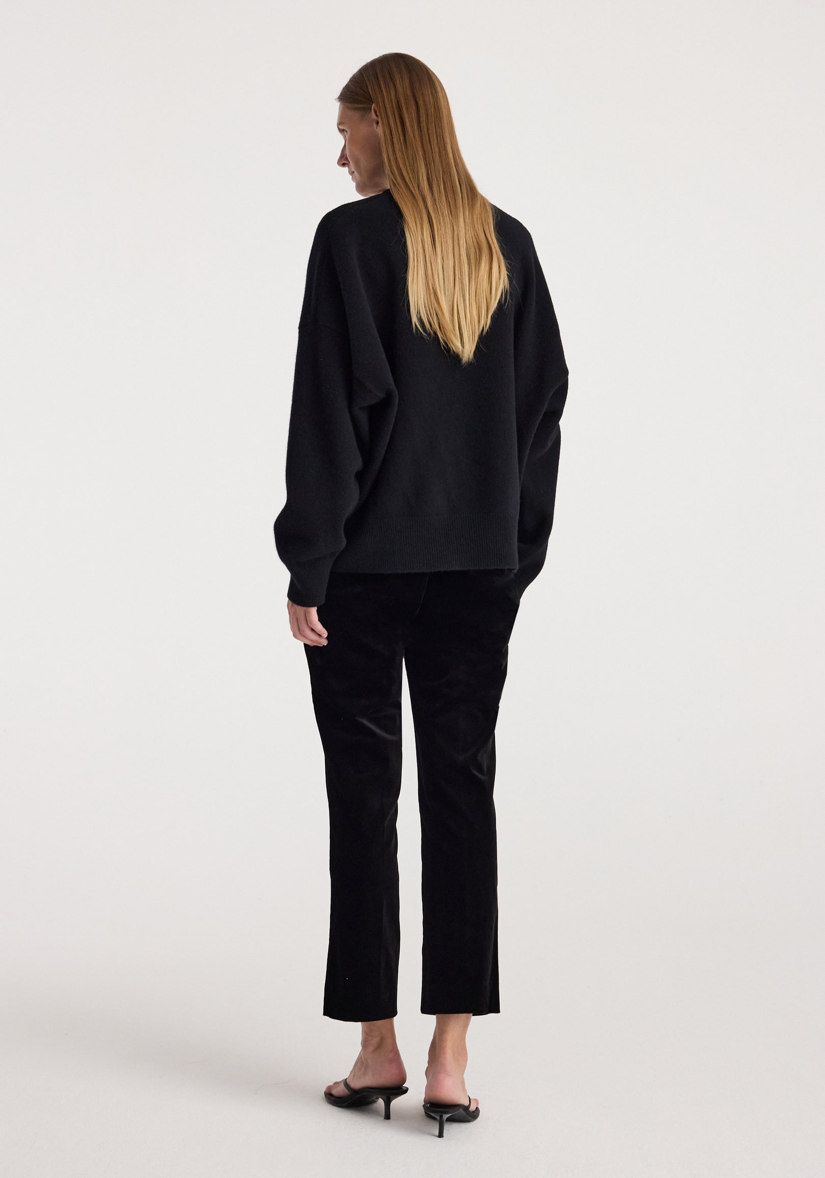 Woman wearing black cigarette leg velvet tailored trousers | black paired with a loose black sweater and black heeled sandals, standing with her back facing the camera.