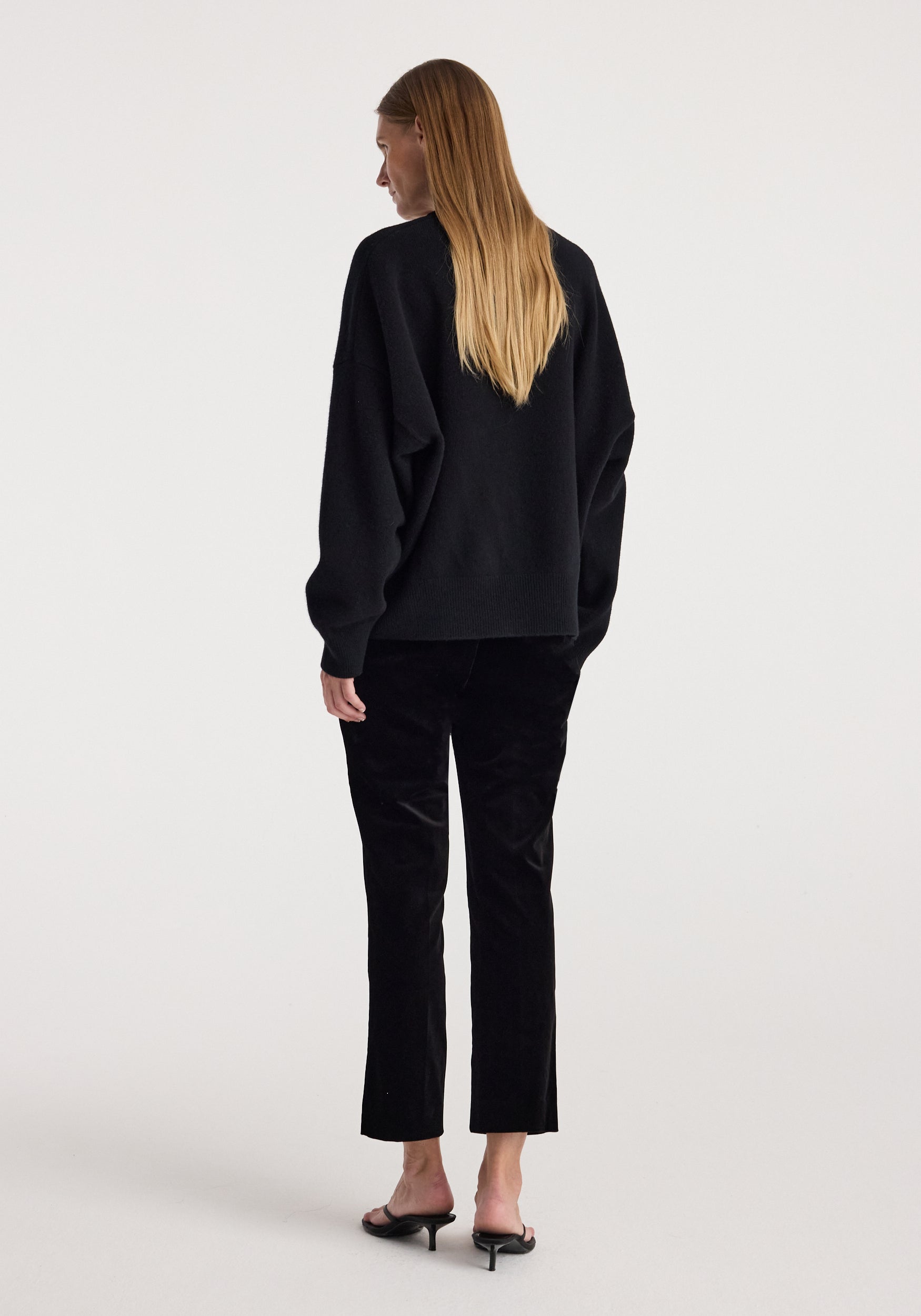 Woman wearing black cigarette leg velvet tailored trousers | black paired with a loose black sweater and black heeled sandals, standing with her back facing the camera.
