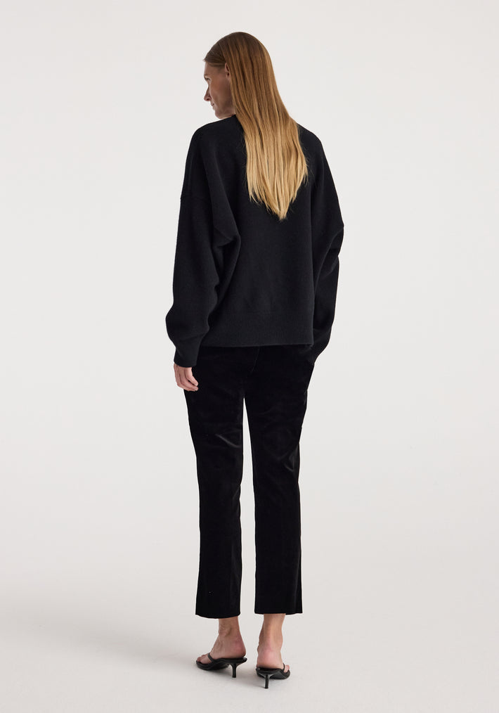 Woman wearing black cigarette leg velvet tailored trousers | black paired with a loose black sweater and black heeled sandals, standing with her back facing the camera.