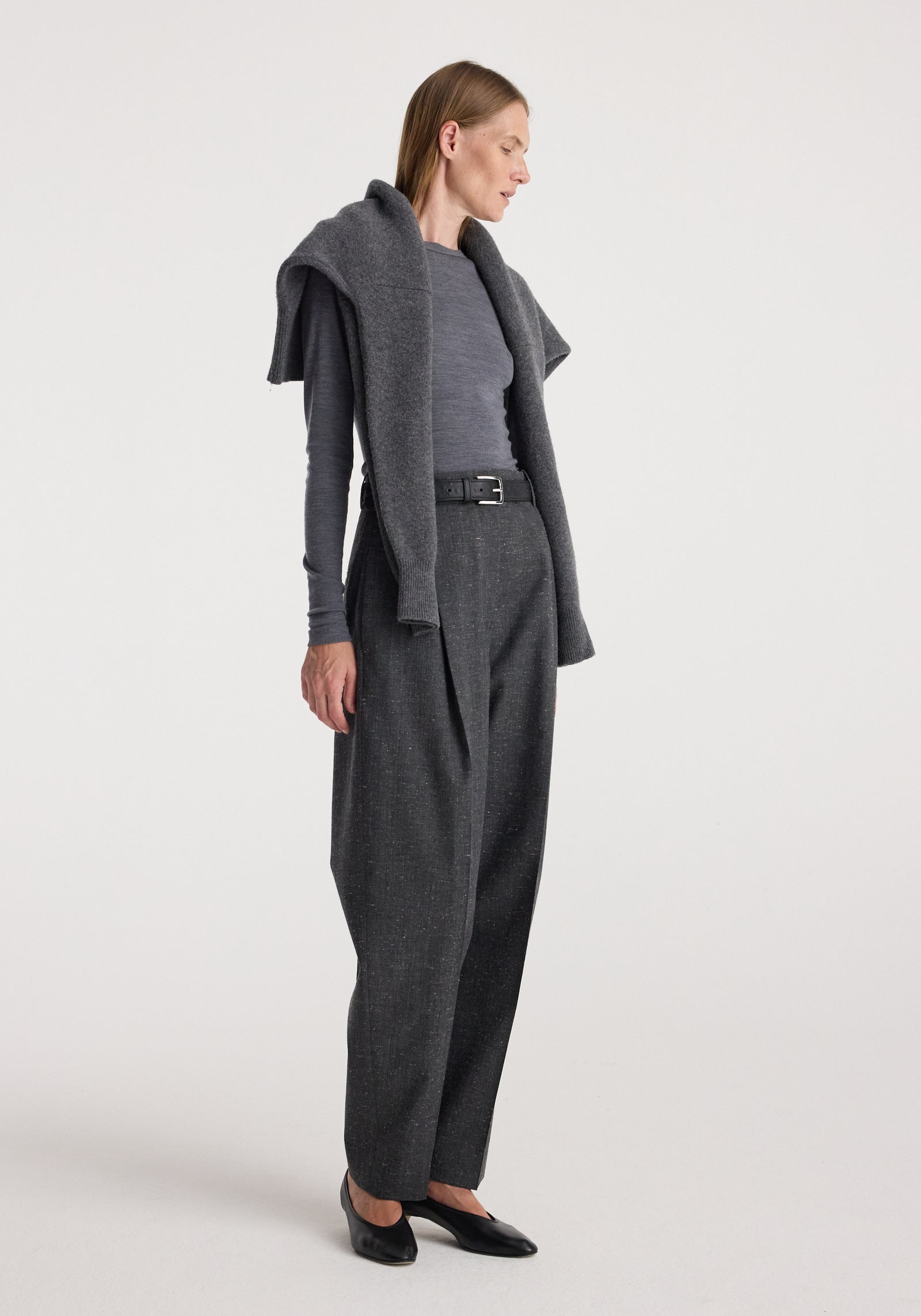Woman wearing Daria - Single pleat straight leg trousers | grey slub paired with a fitted long-sleeve top and a sweater draped over her shoulders, styled with black flats.