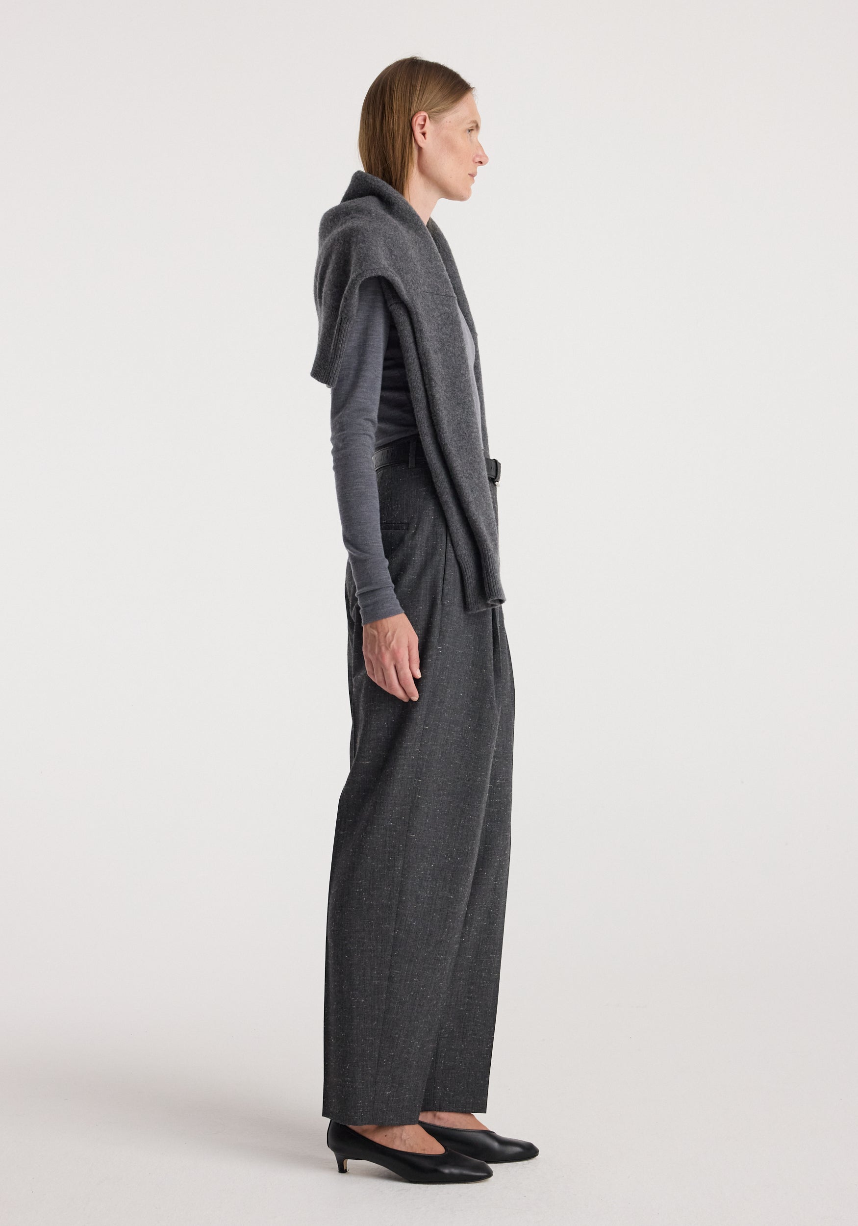 Side profile of a woman wearing Daria - Single pleat straight leg trousers | grey slub paired with a grey long-sleeve top, draped sweater, and black low-heel shoes against a plain background.