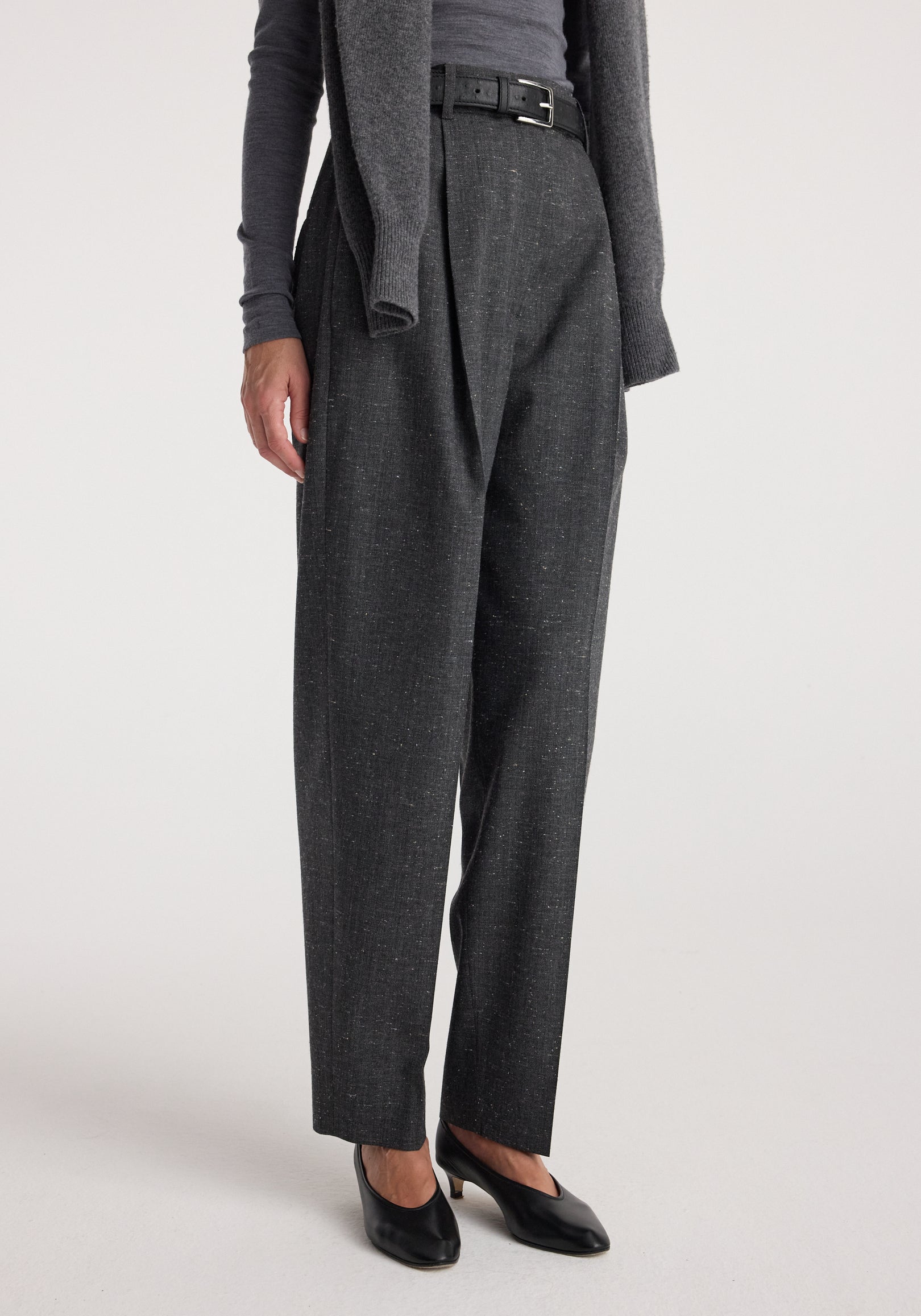 Close-up of a person wearing Daria - Single pleat straight leg trousers | grey slub, styled with a belt, dark top, and black low-heeled shoes against a plain background.