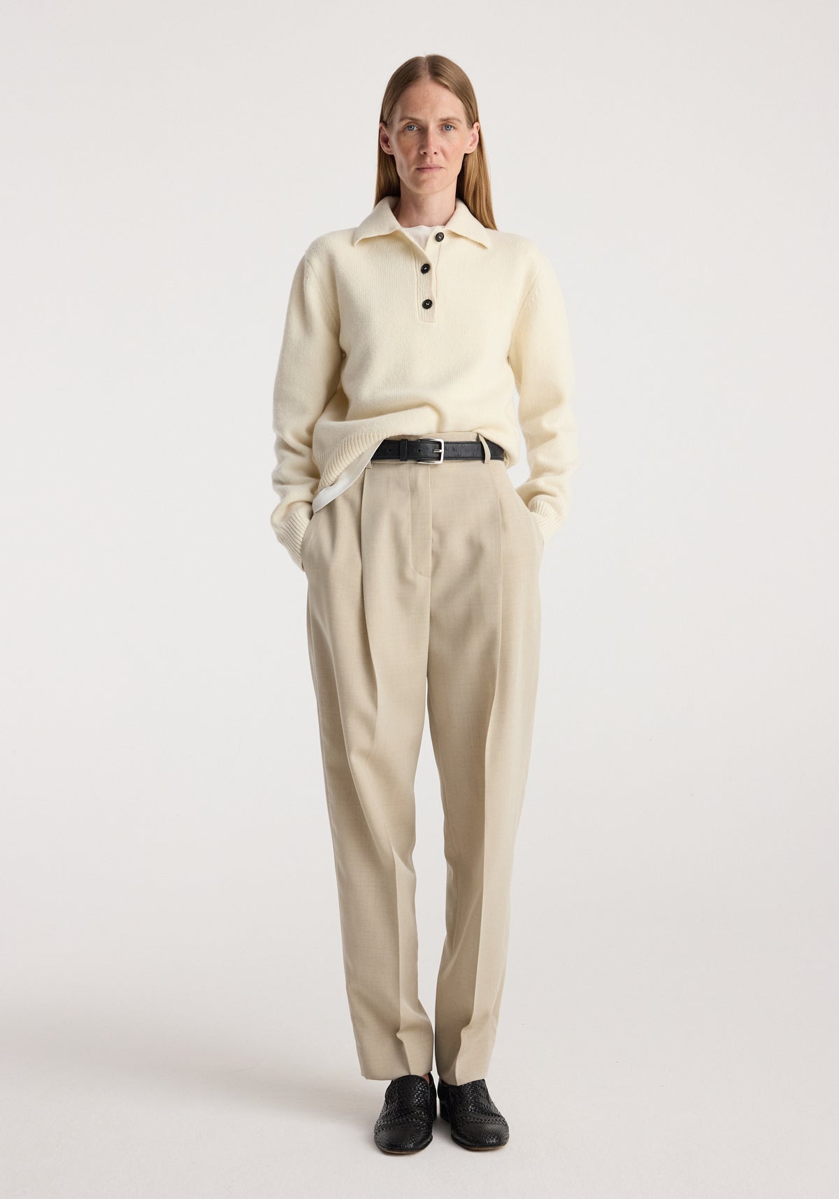 Woman wearing cream knit sweater and Daria - Single pleat straight leg trousers | butter, styled with black belt and black shoes, standing against plain light background.
