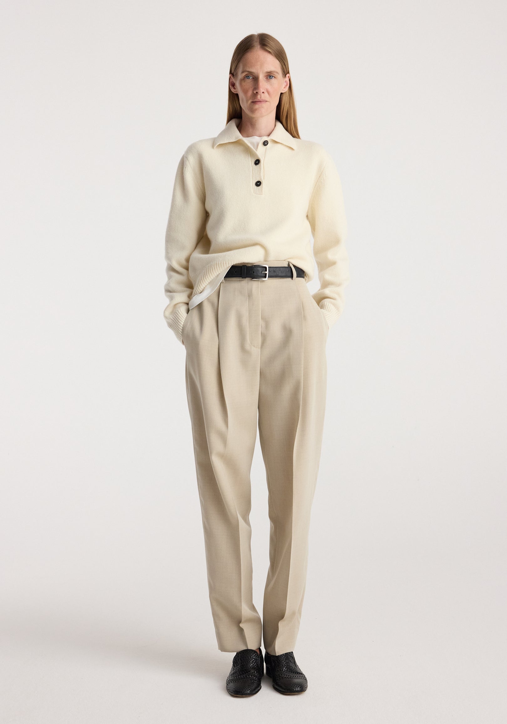 Woman wearing cream knit sweater and Daria - Single pleat straight leg trousers | butter, styled with black belt and black shoes, standing against plain light background.