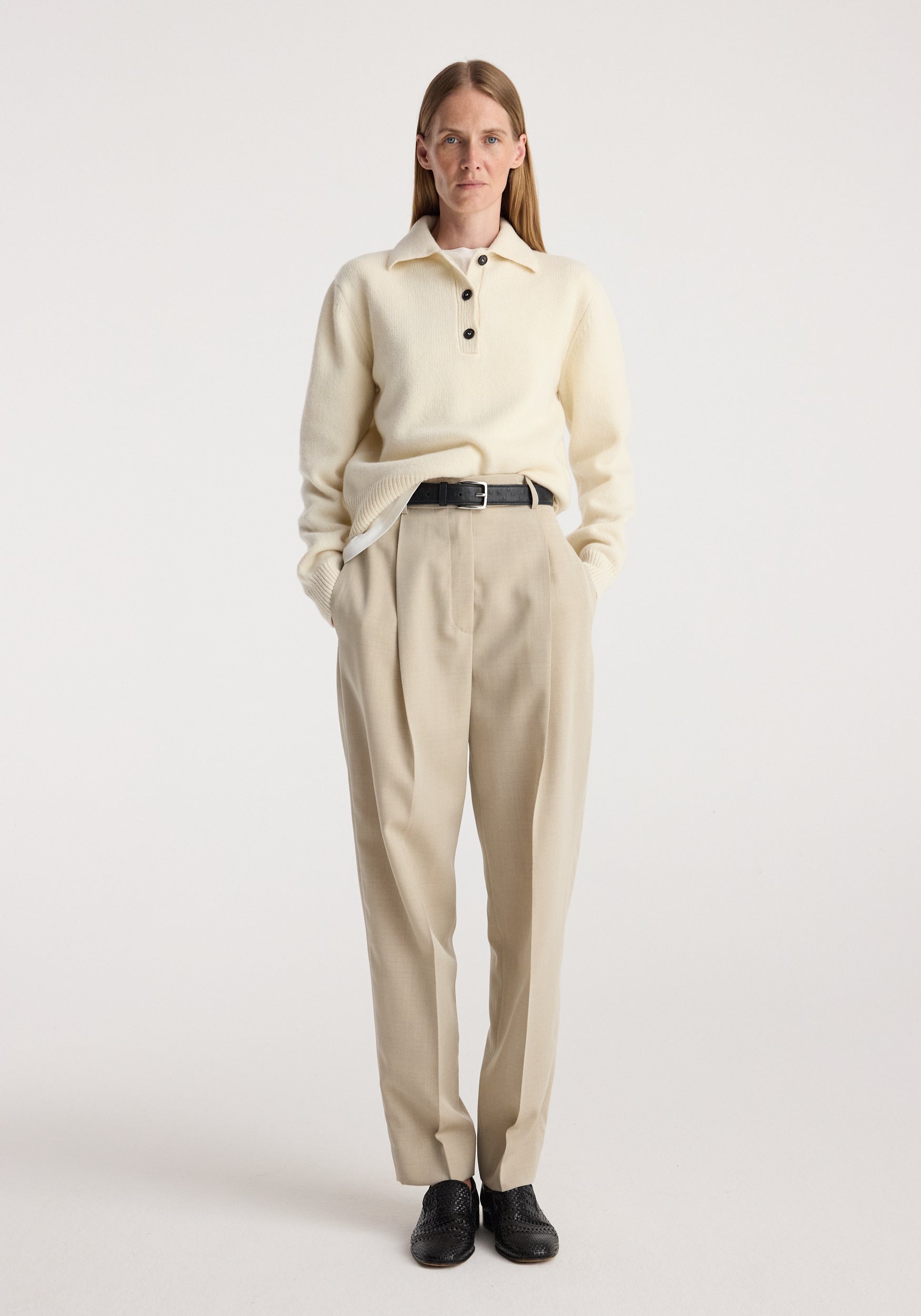 Woman wearing cream knit sweater and Daria - Single pleat straight leg trousers | butter, styled with black belt and black shoes, standing against plain light background.