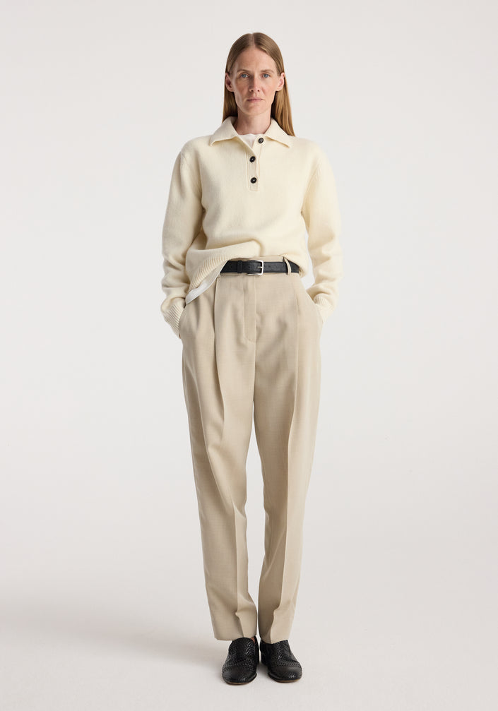 Woman wearing cream knit sweater and Daria - Single pleat straight leg trousers | butter, styled with black belt and black shoes, standing against plain light background.