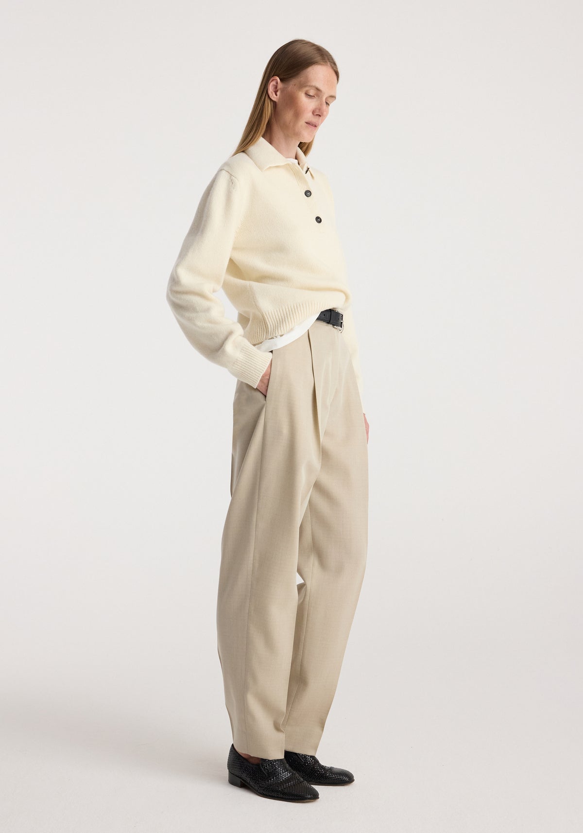 Woman wearing Daria - Single pleat straight leg trousers | butter in a neutral beige shade paired with a cream sweater and black shoes standing against a plain background.