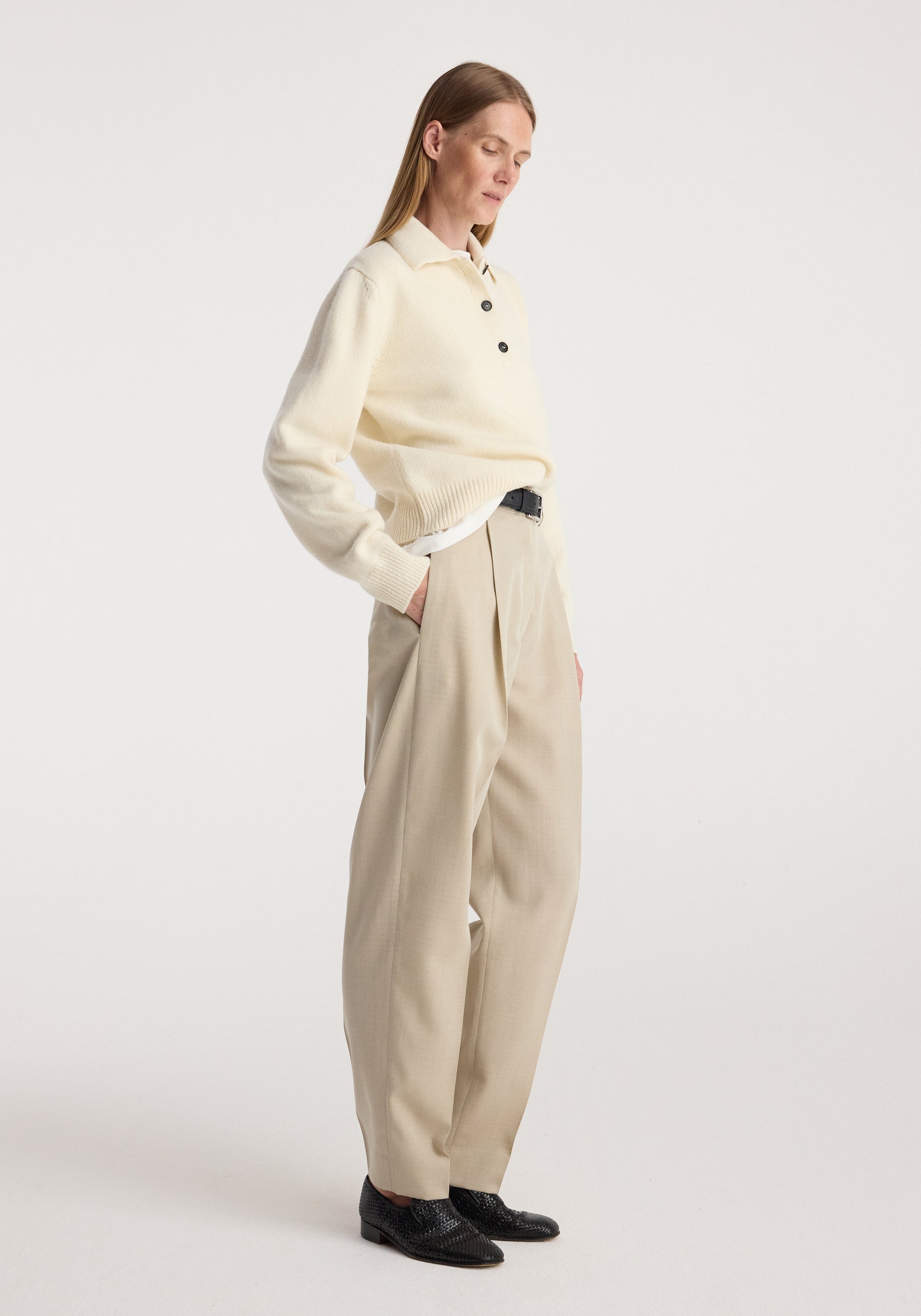 Woman wearing Daria - Single pleat straight leg trousers | butter in a neutral beige shade paired with a cream sweater and black shoes standing against a plain background.