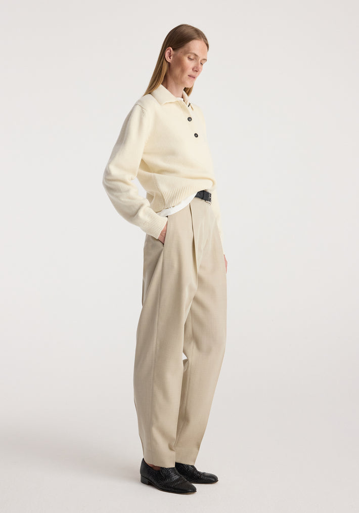 Woman wearing Daria - Single pleat straight leg trousers | butter in a neutral beige shade paired with a cream sweater and black shoes standing against a plain background.