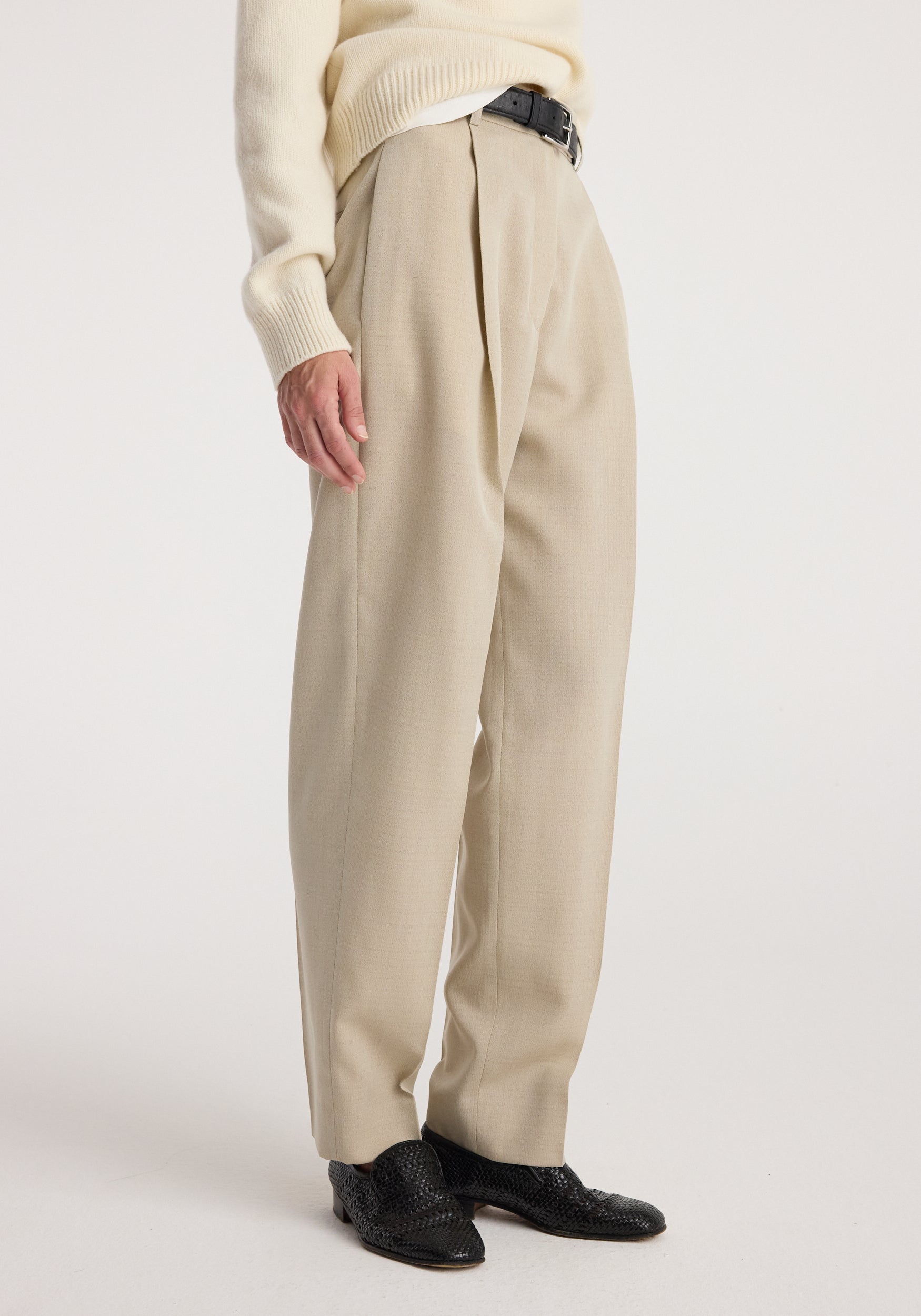 Side view of a person wearing Daria - Single pleat straight leg trousers | butter styled with a cream sweater, black belt, and black textured shoes against a white background.