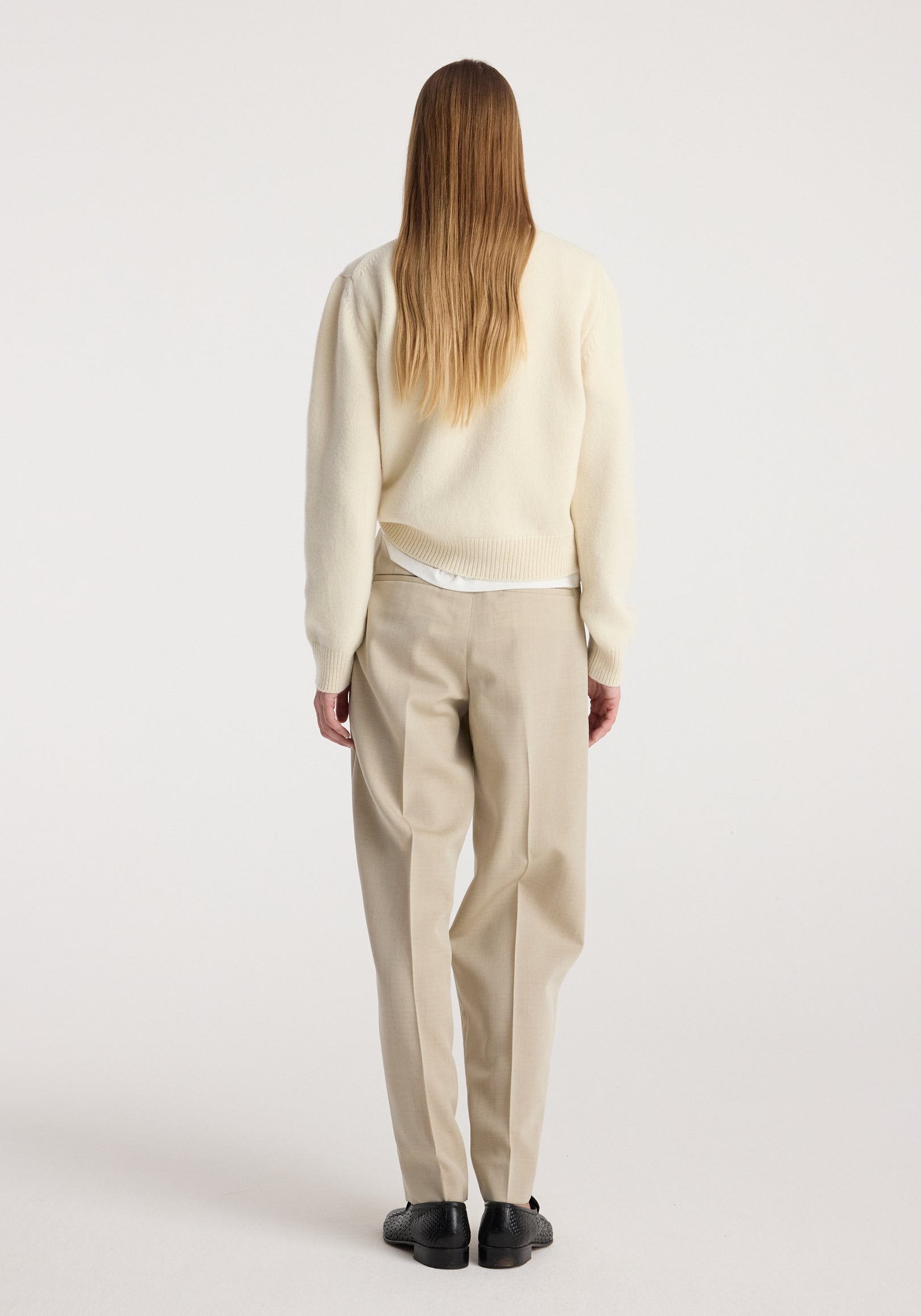 Woman seen from the back wearing Daria - Single pleat straight leg trousers | butter in a light beige shade, paired with a cream sweater and black shoes against a plain background.