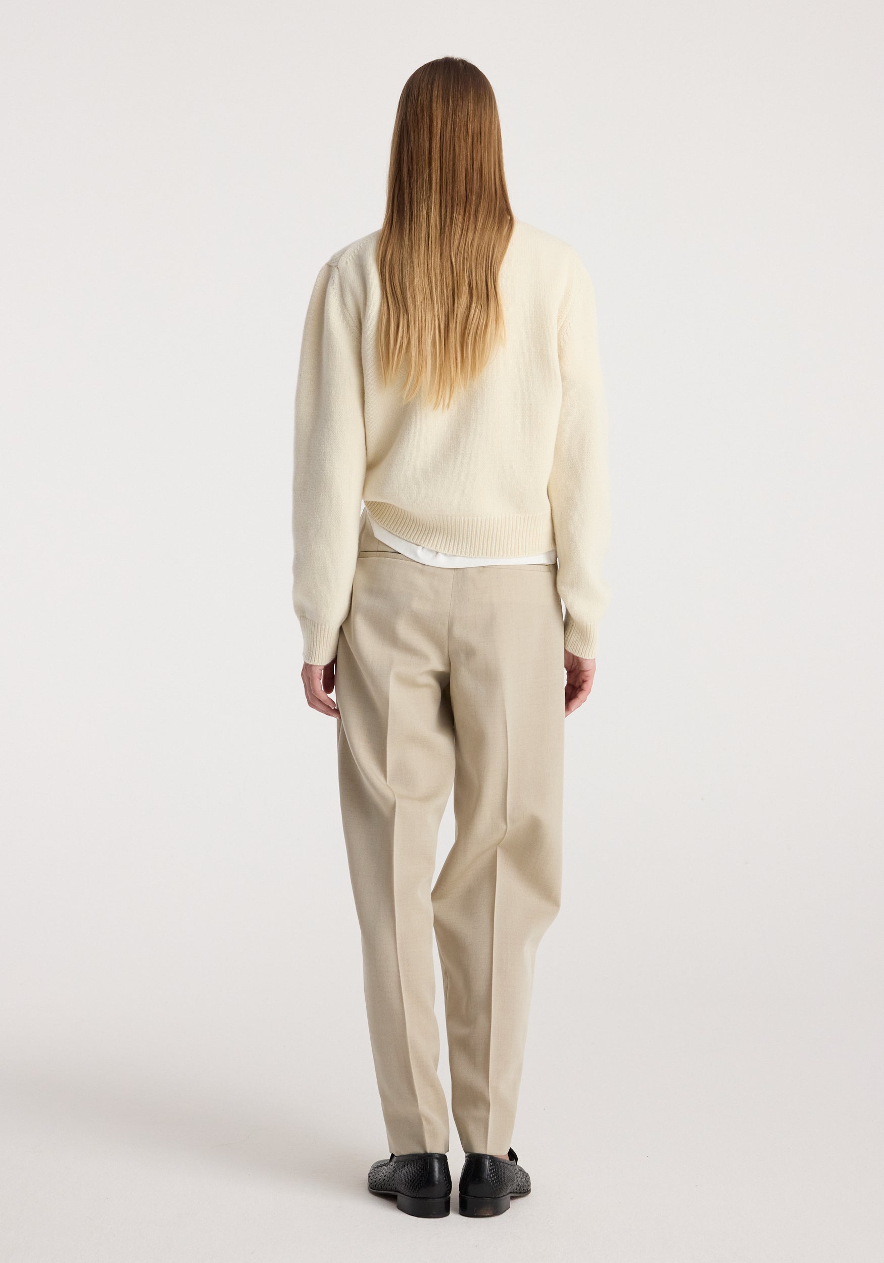 Woman seen from the back wearing Daria - Single pleat straight leg trousers | butter in a light beige shade, paired with a cream sweater and black shoes against a plain background.