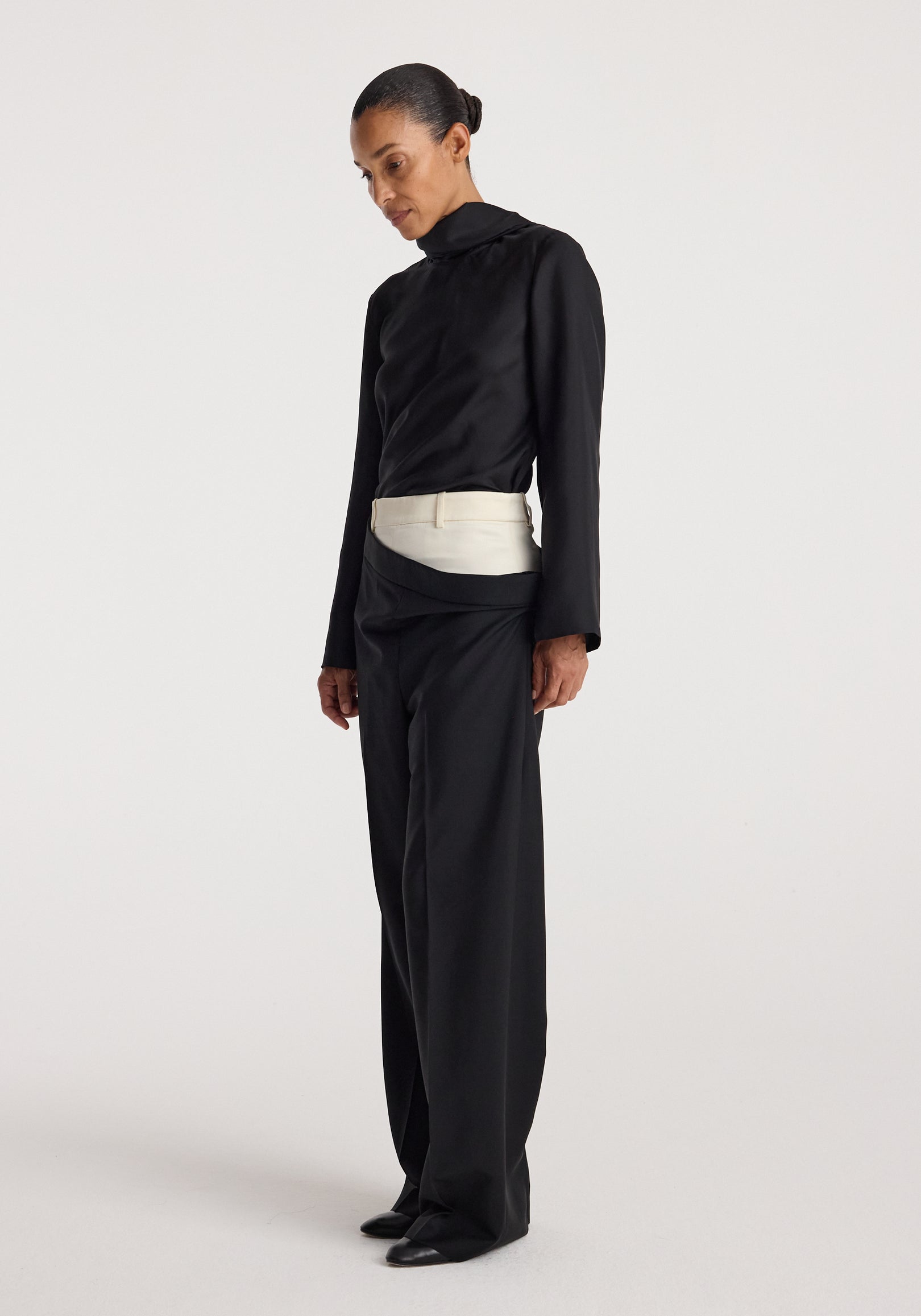 Model wearing Double waistband trousers | black with a contrasting light-colored double waistband detail, paired with a black long-sleeve top and black shoes, standing against a plain background.