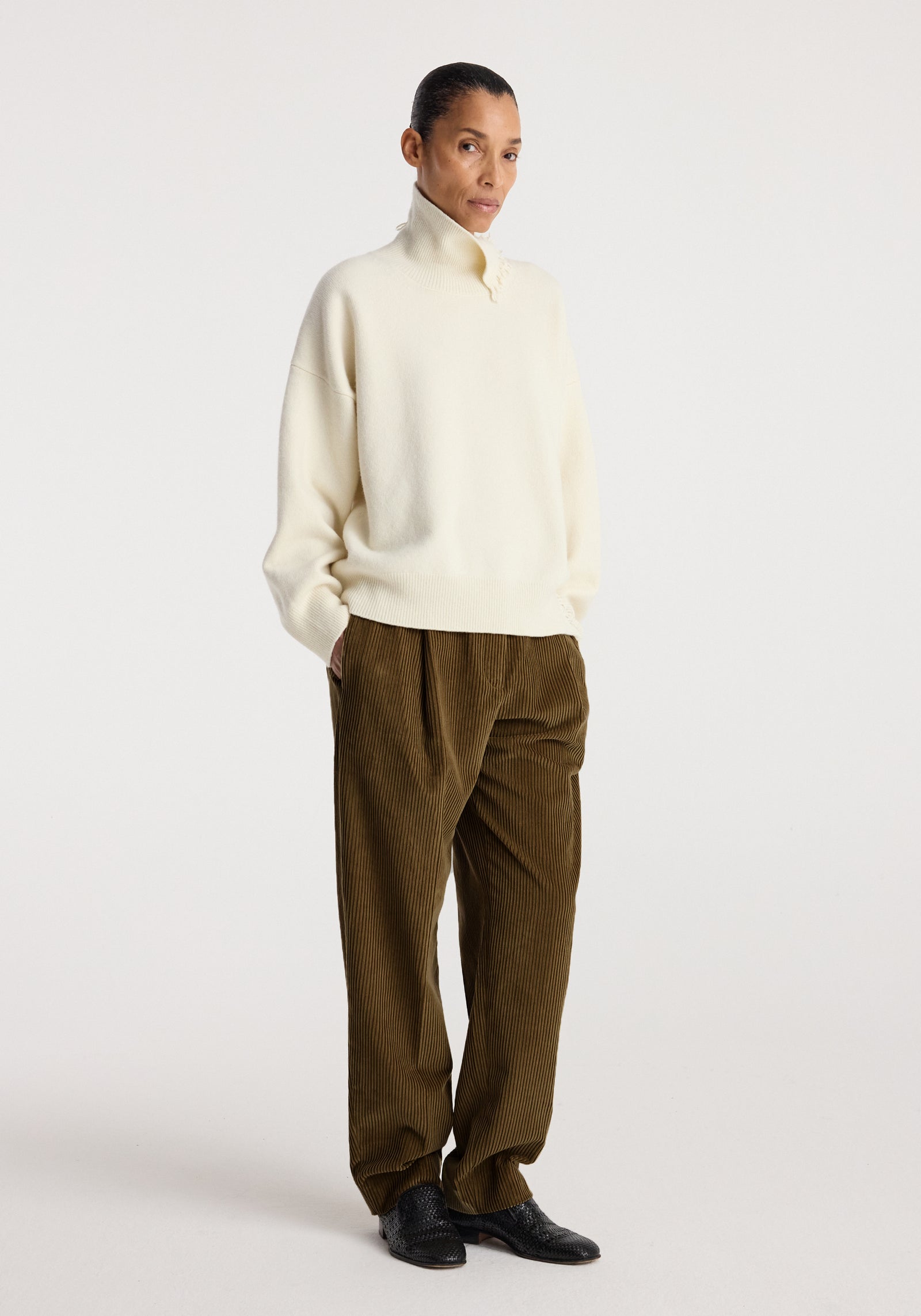 Model wearing Daria - Single pleat straight leg corduroy trousers | caramel paired with an oversized cream sweater and black shoes against a plain background.