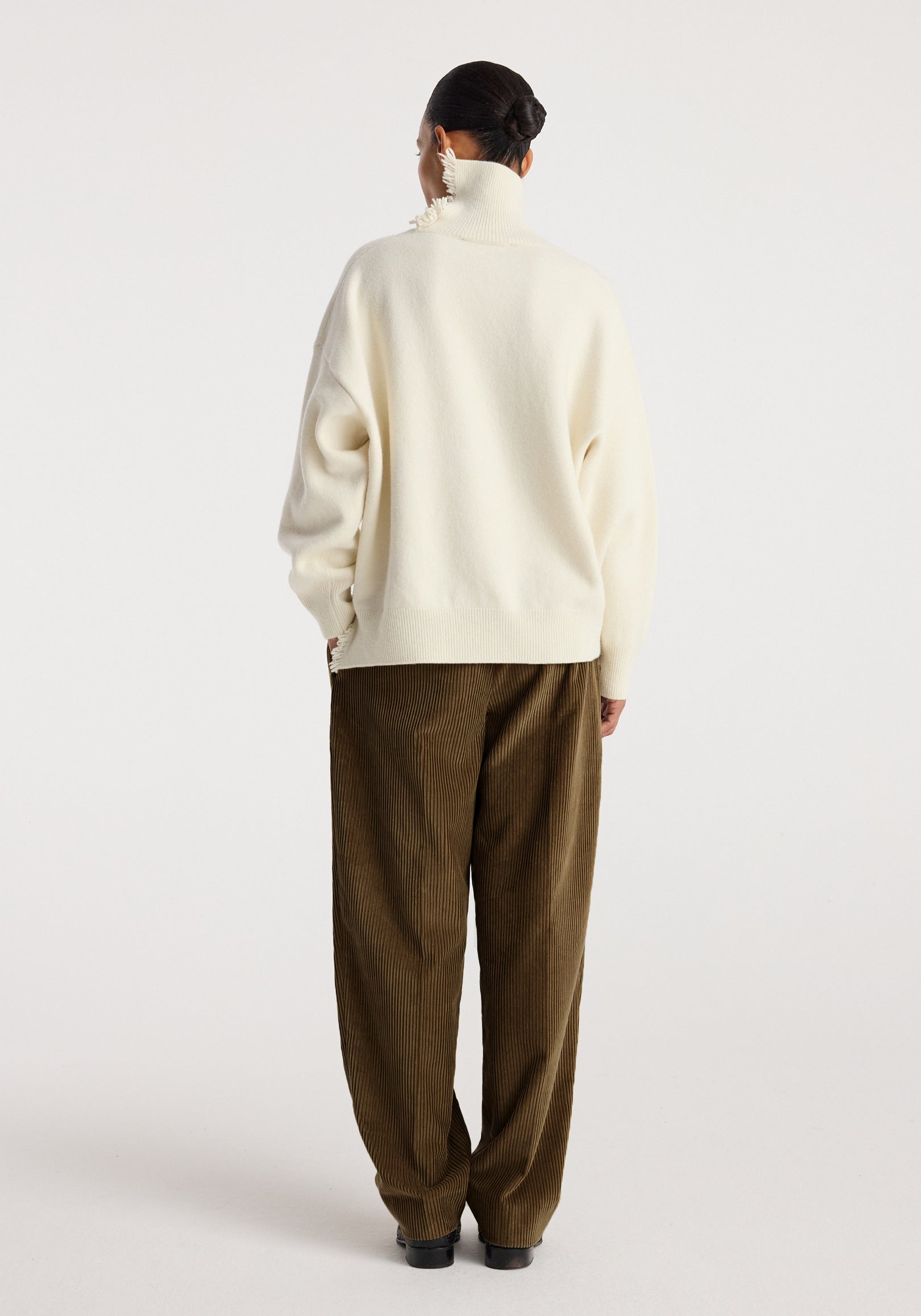 Back view of a woman wearing Daria - Single pleat straight leg corduroy trousers | caramel with a white oversized turtleneck sweater and black shoes against a plain background.