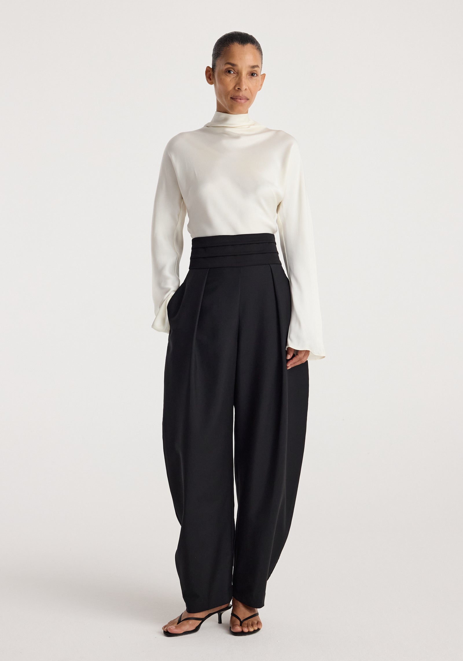 Woman wearing a white long-sleeve top paired with black Puck - Sculptural cumberband trousers | black, featuring a high waist and pleated front design.