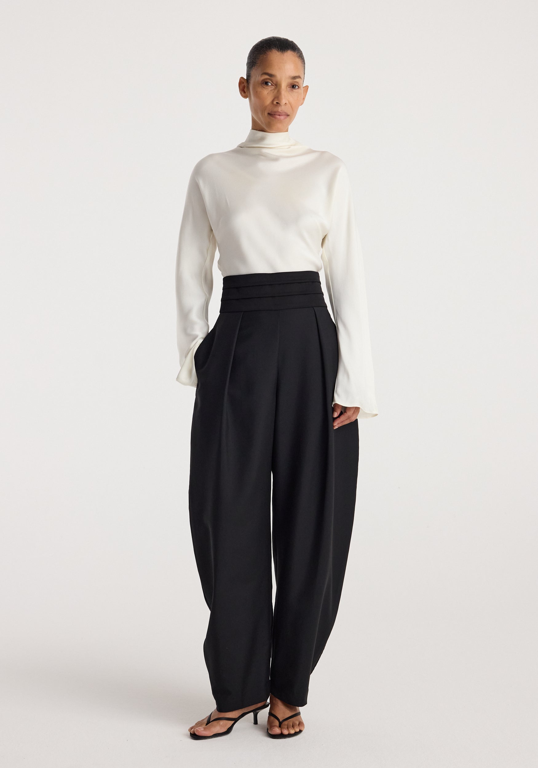 Woman wearing a white long-sleeve top paired with black Puck - Sculptural cumberband trousers | black, featuring a high waist and pleated front design.