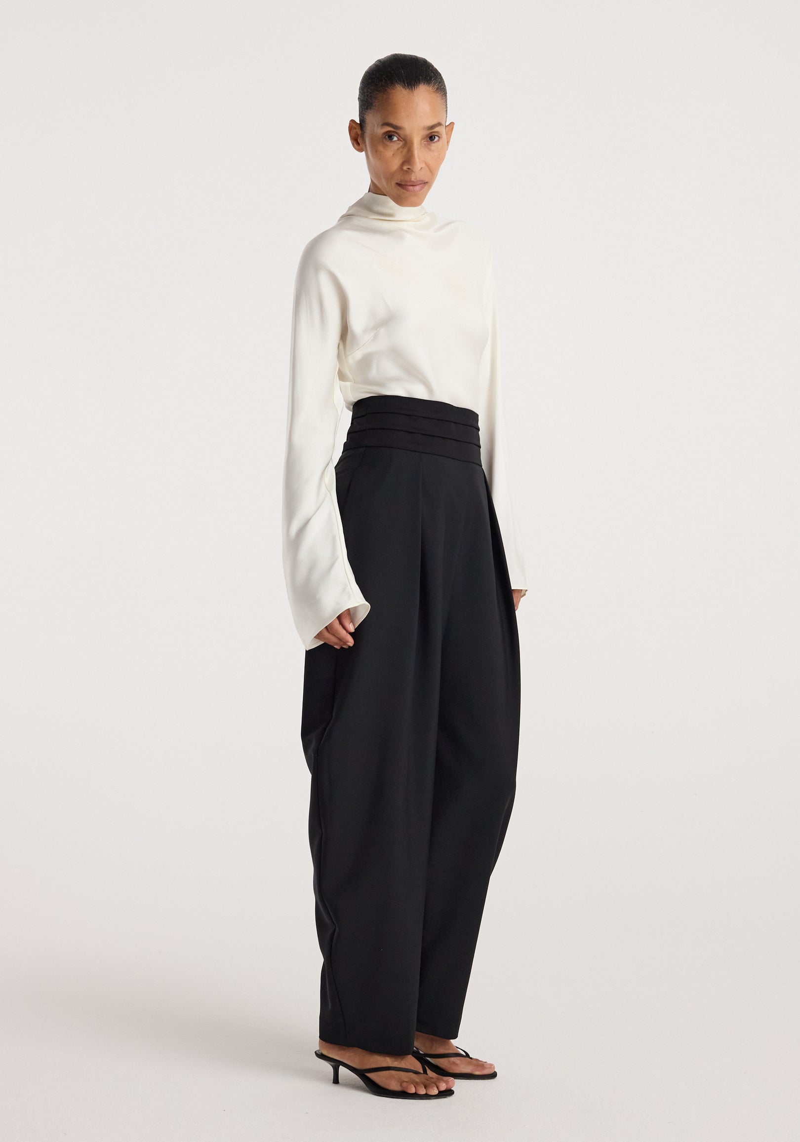 Woman wearing black Puck - Sculptural cumberband trousers | black paired with a white long-sleeve top and black heeled sandals standing against a plain background.