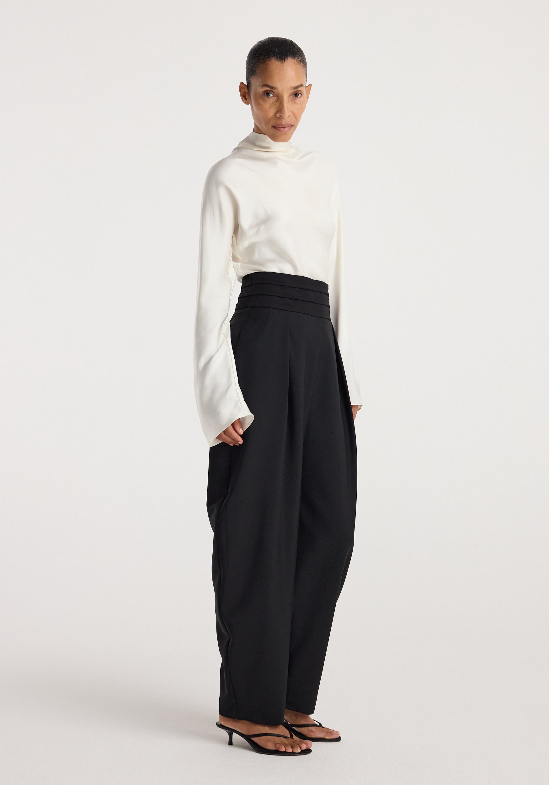 Woman wearing black Puck - Sculptural cumberband trousers | black paired with a white long-sleeve top and black heeled sandals standing against a plain background.