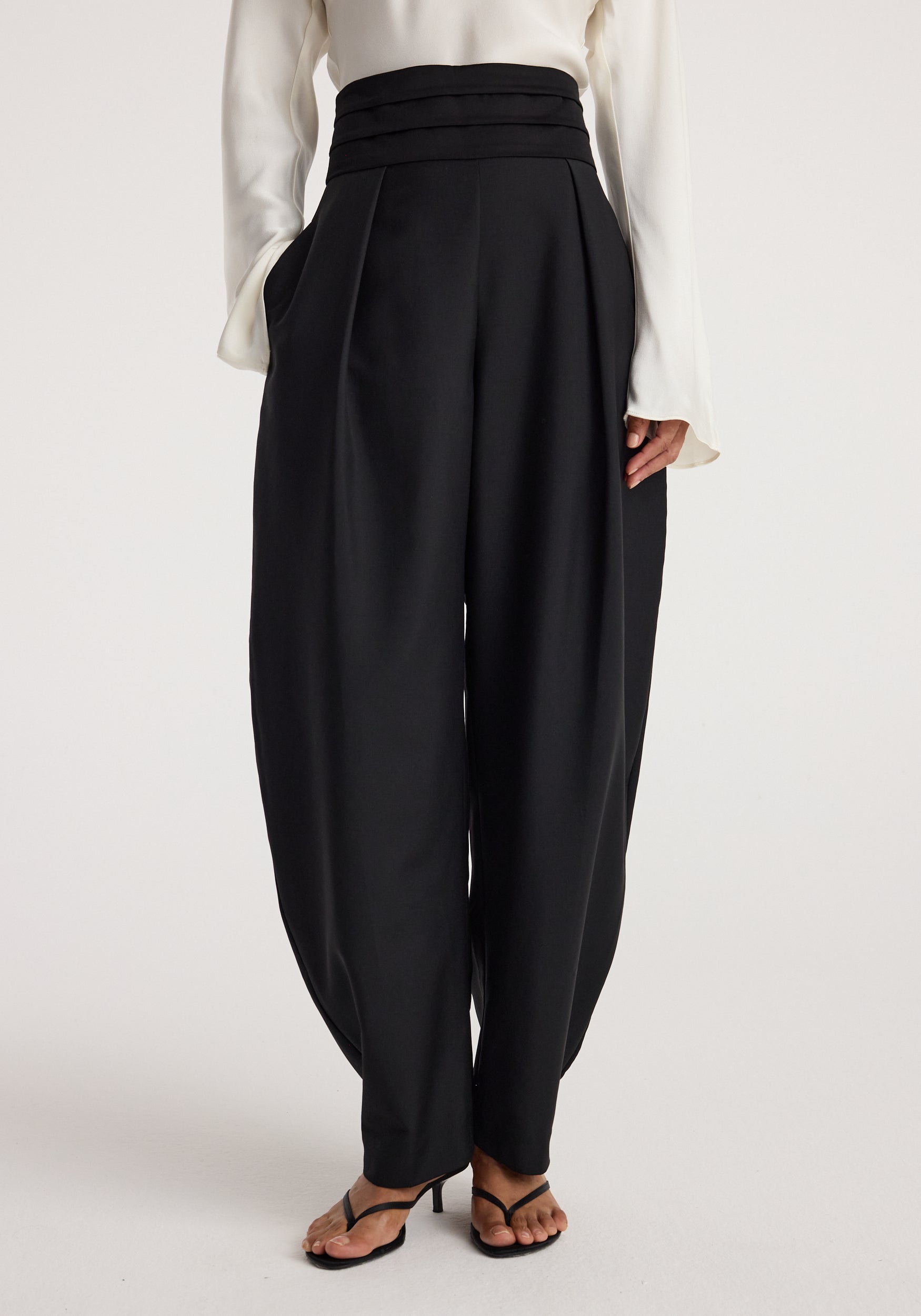Front view of a model wearing black "Puck - Sculptural cumberband trousers | black" with a high, structured waistband, wide tapered legs, and side pockets paired with black heeled sandals.
