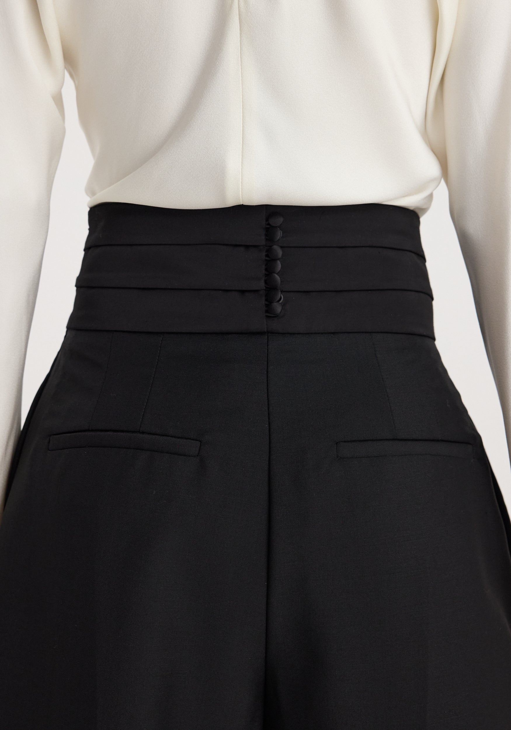 Back view of a person wearing black Puck - Sculptural cumberband trousers | black with a high waist and buttoned structured waistband paired with a white top.