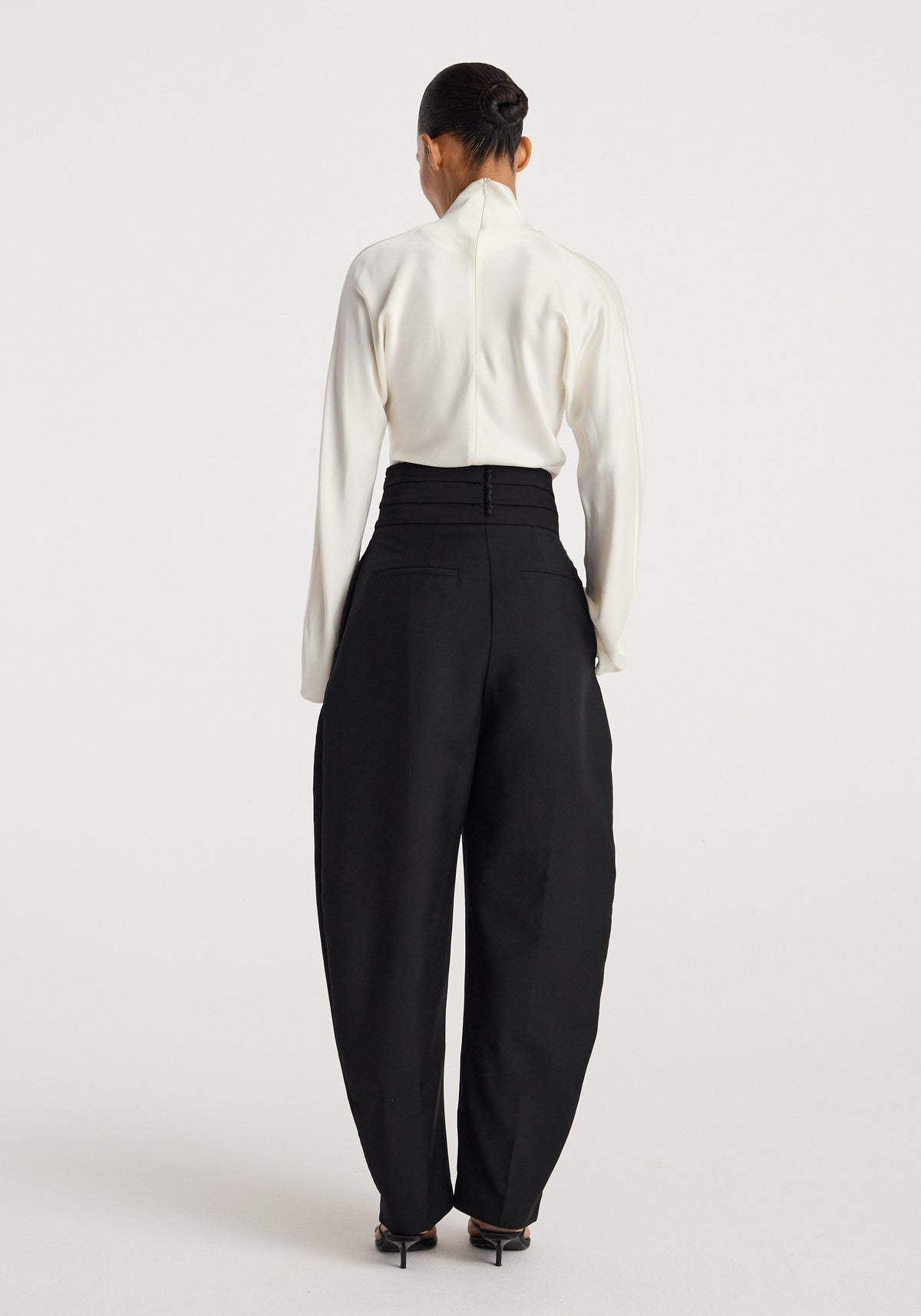 Woman wearing black Puck - Sculptural cumberband trousers | black paired with a white long-sleeve top, shown from the back in a minimal studio setting.