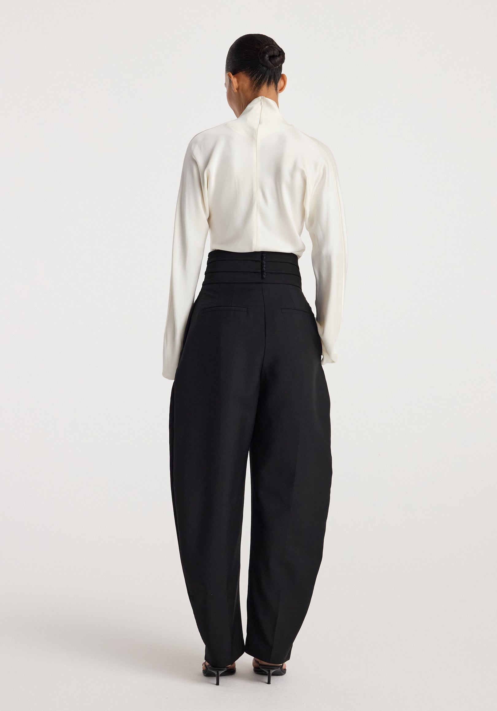 Woman wearing black Puck - Sculptural cumberband trousers | black paired with a white long-sleeve top, shown from the back in a minimal studio setting.