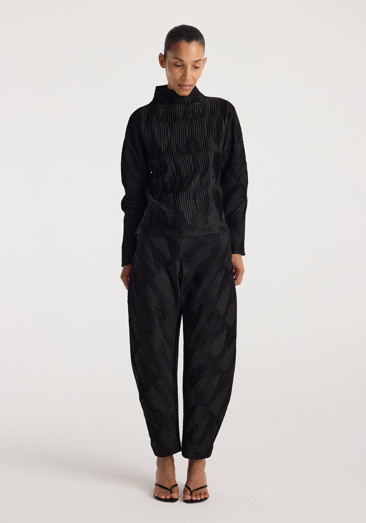 Woman wearing black Plissé satin trousers | black with a matching long-sleeve top, standing against a plain light background and looking downward.